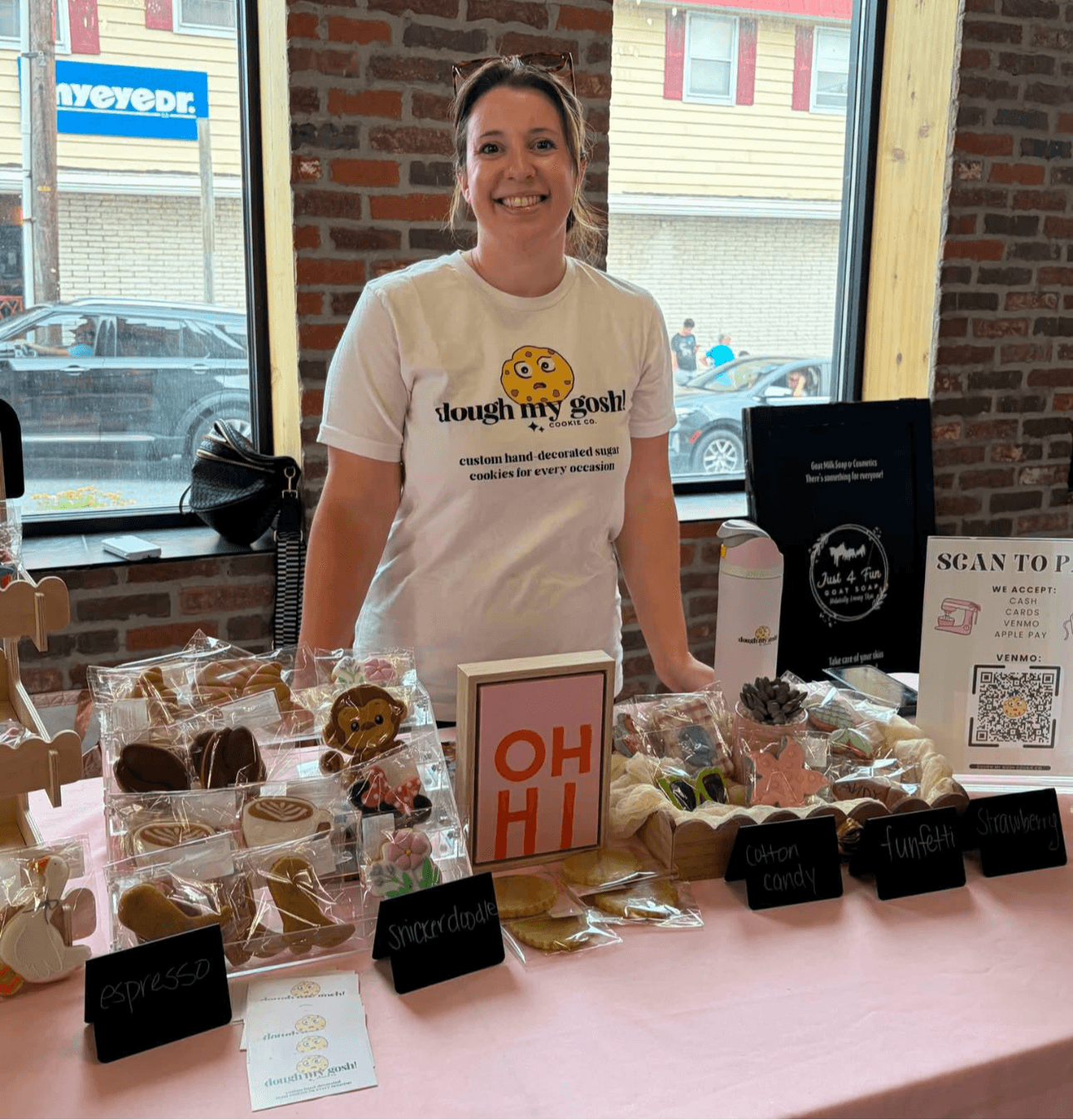 Woman standing behind a table with baked goods, smiling, in front of a window with brick wall outside.