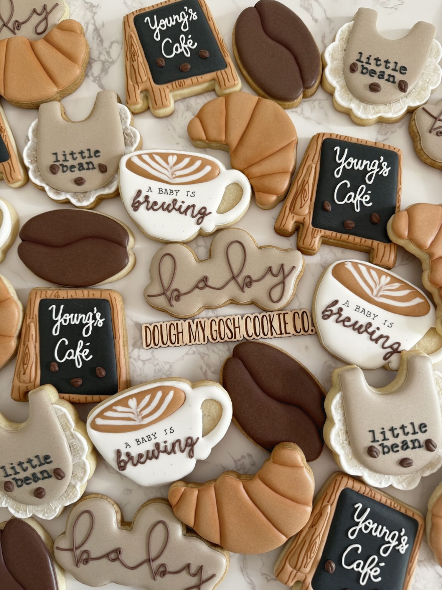Assorted baby-themed cookies with text, coffee cups, and baby items, decorated in brown, white, and beige colors.