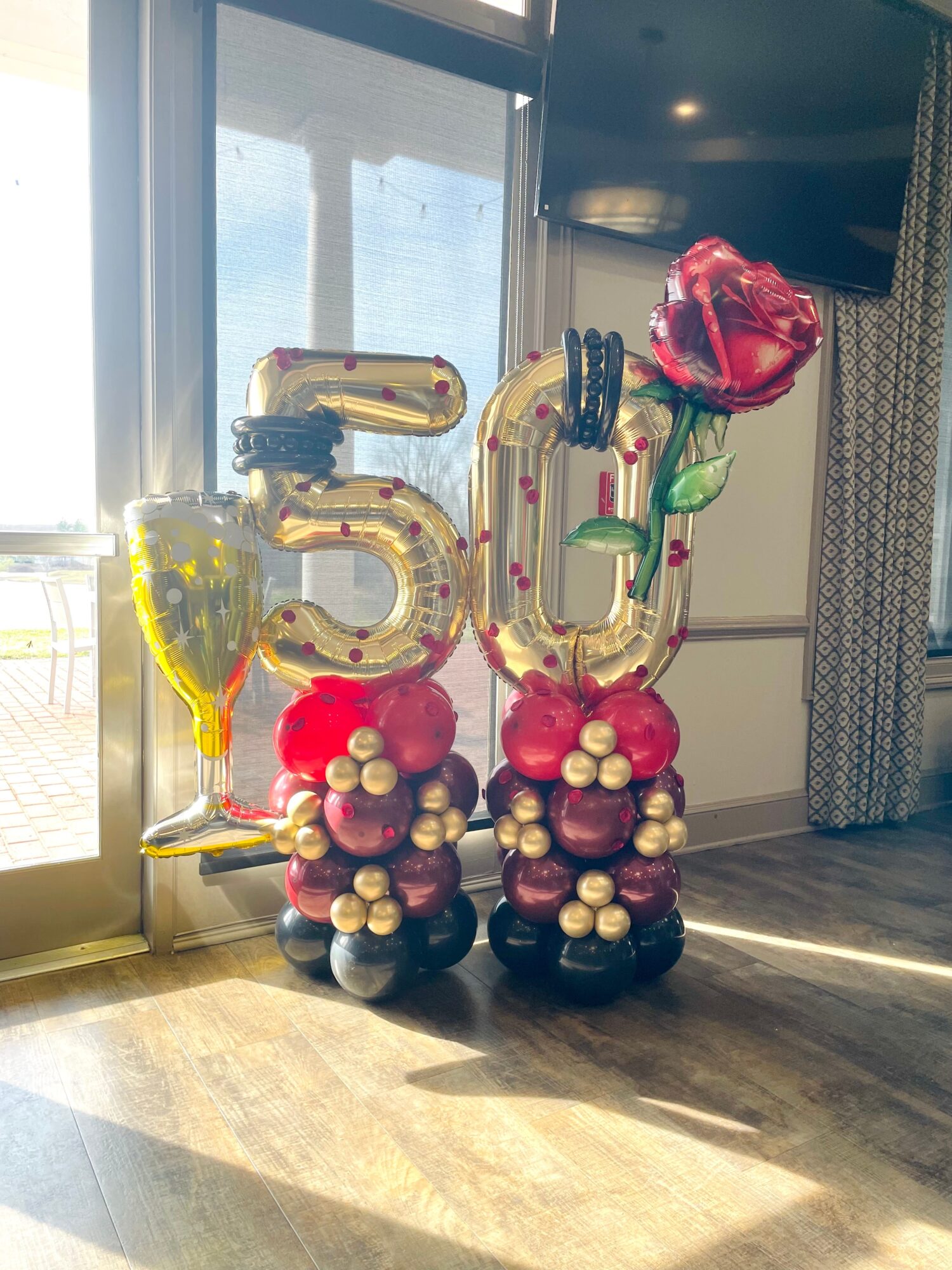 Celebration balloons with the number 50, a champagne glass, and a rose, arranged on a wooden floor near a window.