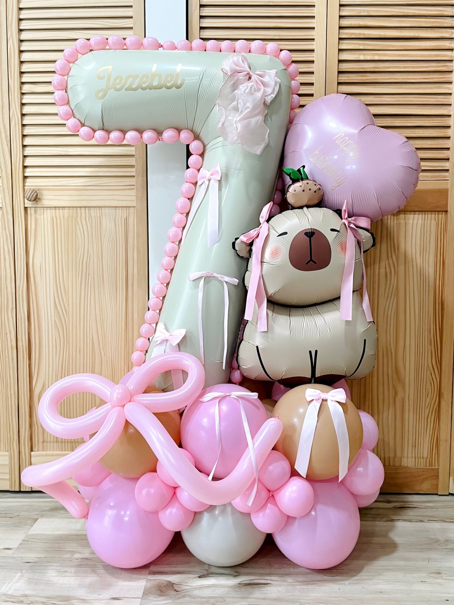 Decorative balloon arrangement with a large number seven, a bear, and heart-shaped balloons in pink and white, on wooden background.