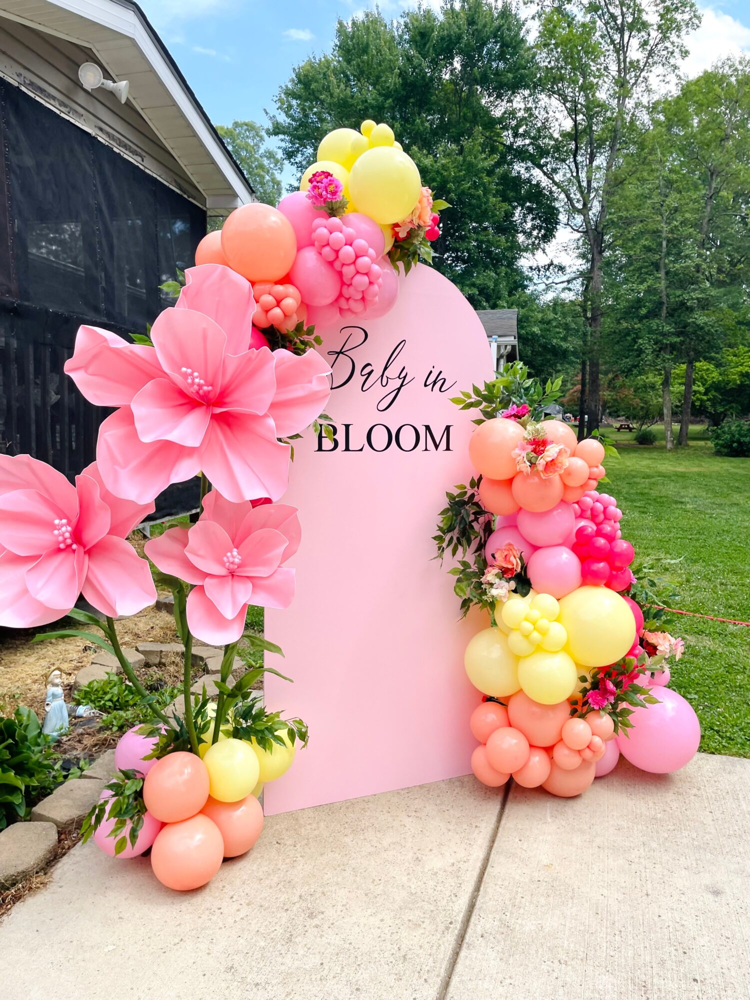 Pink floral arch with balloons and a pink sign reading 'Baby in Bloom' outdoors with trees and grass.