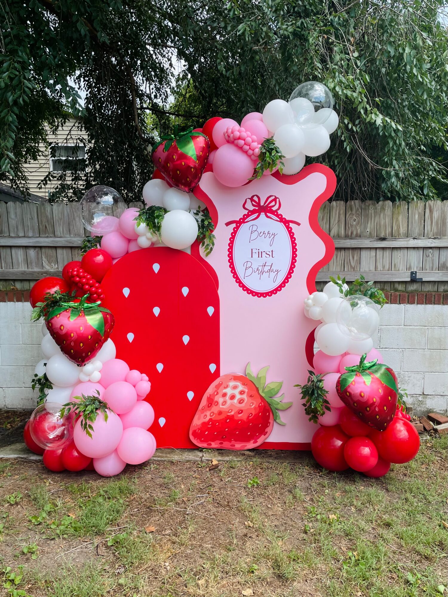 Decorative pink and red balloon arch with strawberries and a pink sign reading 'First First Birthday' outdoors.