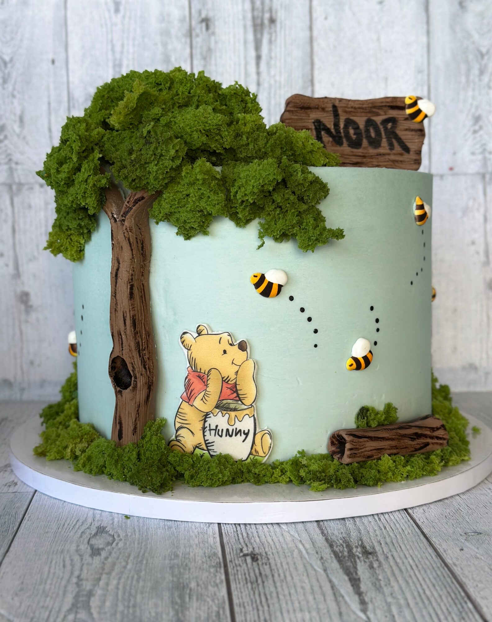 Cake decorated with a tree, bees, a bear holding a honey jar, and a wooden sign that says 'Noor'.