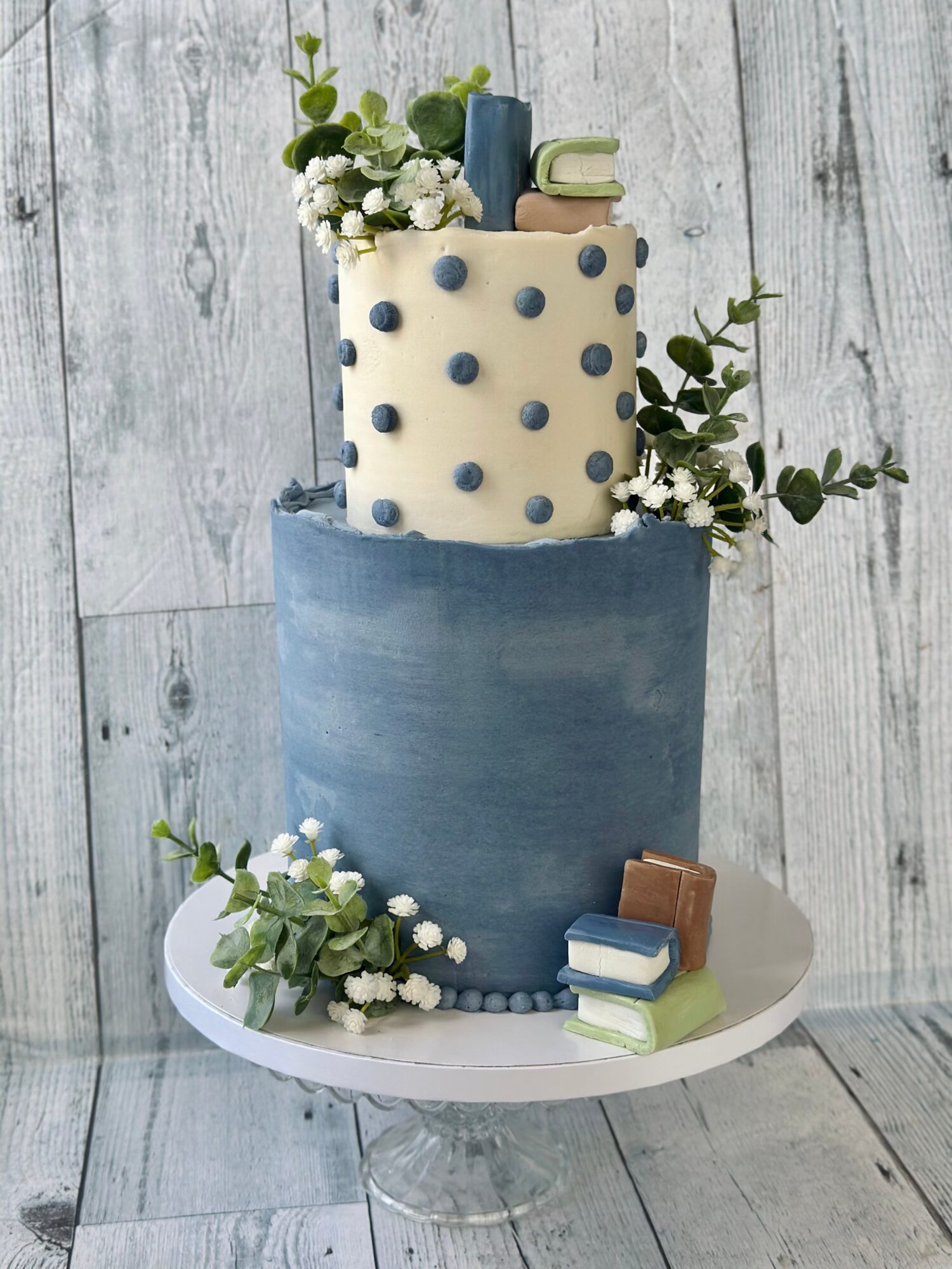 Two-tiered cake with blue and white decorations, topped with flowers and books, on a white stand against a wooden background.