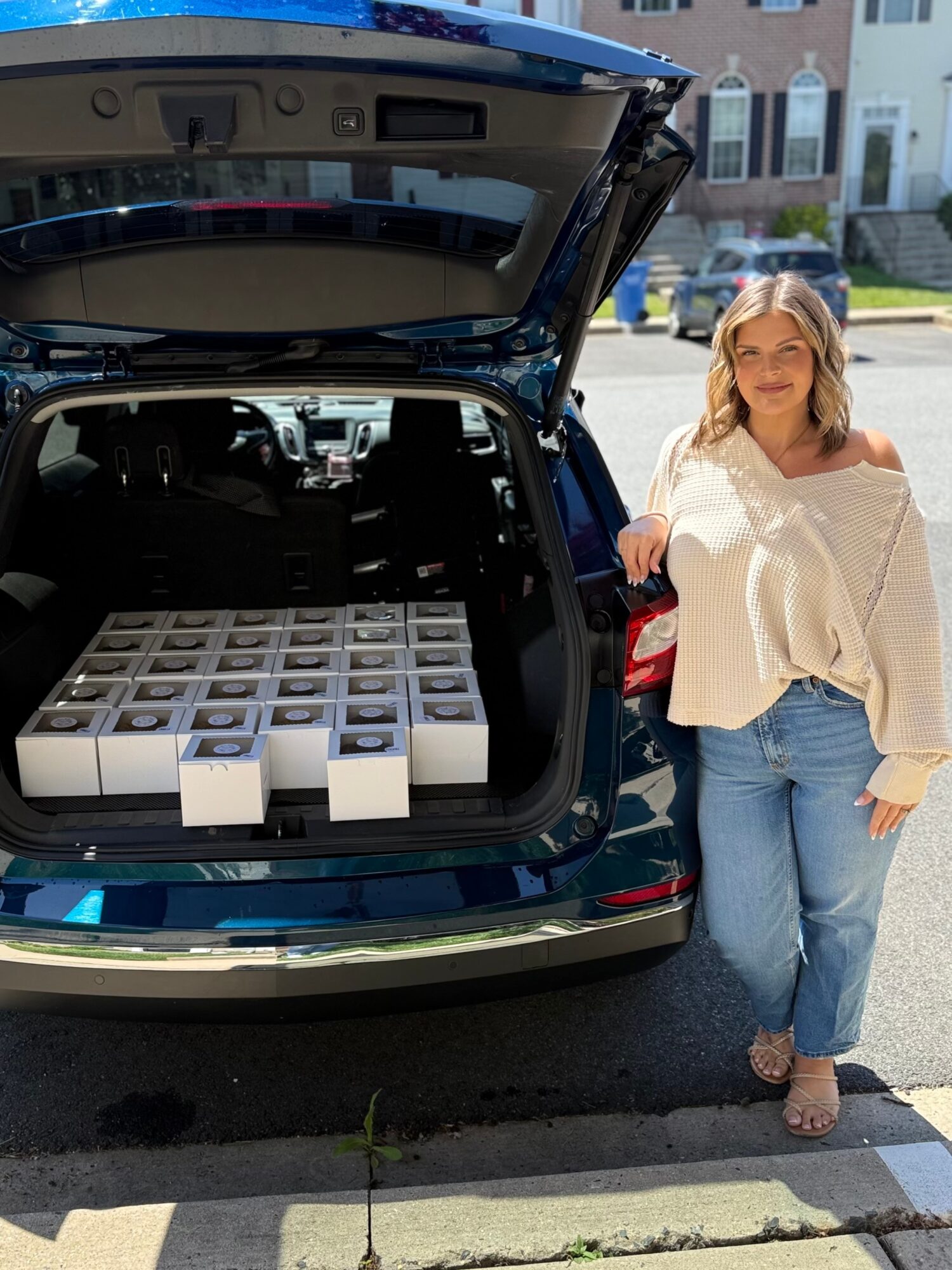 Woman standing next to an open car trunk filled with boxes, smiling outdoors on a sunny day.