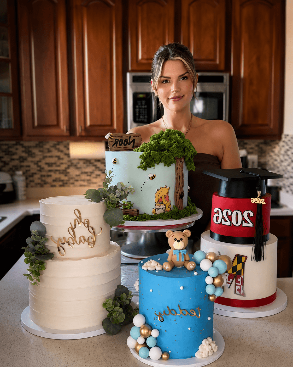 Woman with three decorated cakes in a kitchen, one with a teddy bear and blue theme, another with a lion and greenery, and a third with a graduation cap. 20 words.