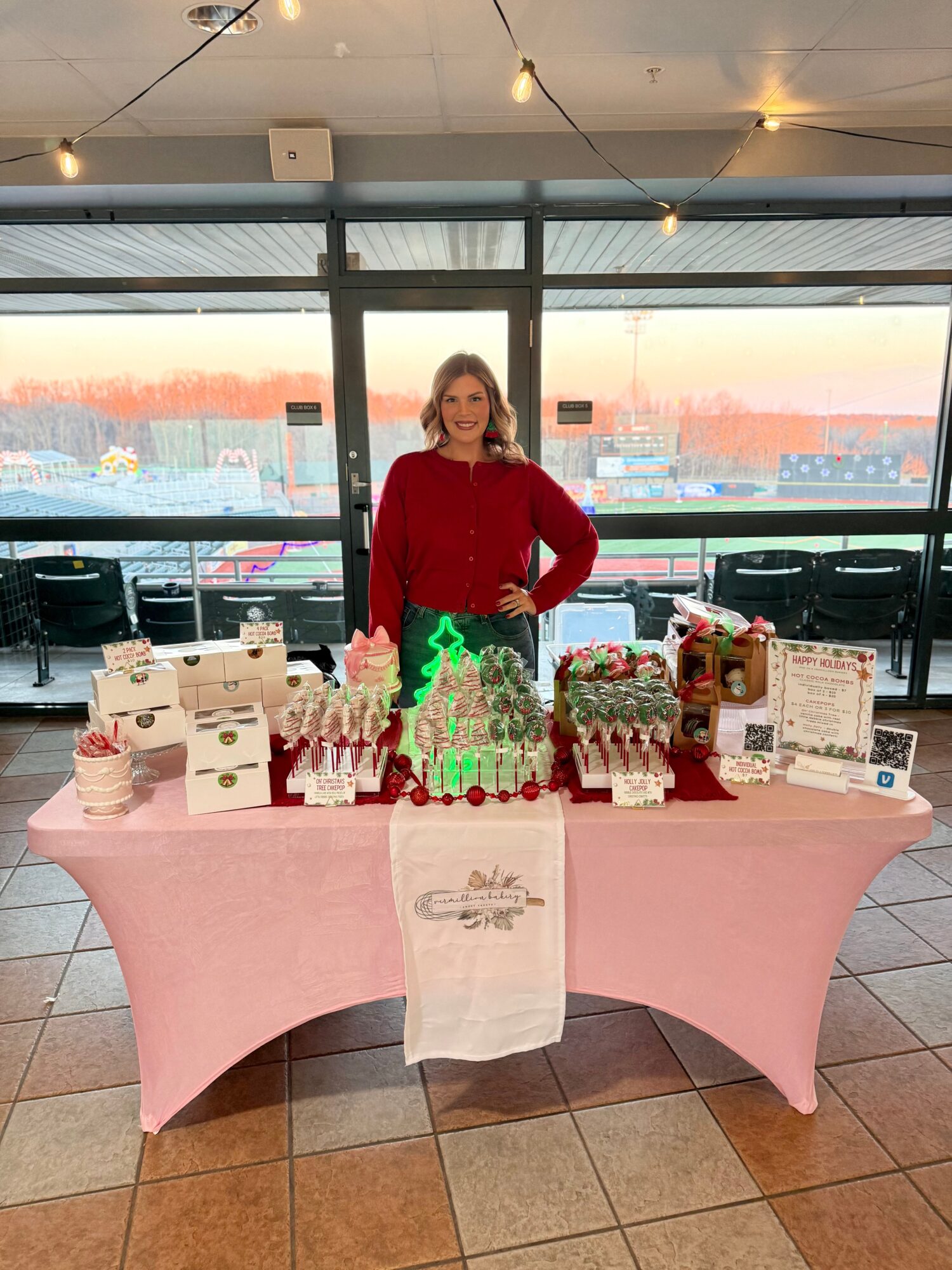 Woman standing behind a table with baked goods and decorations, large windows with a sunset view in the background.