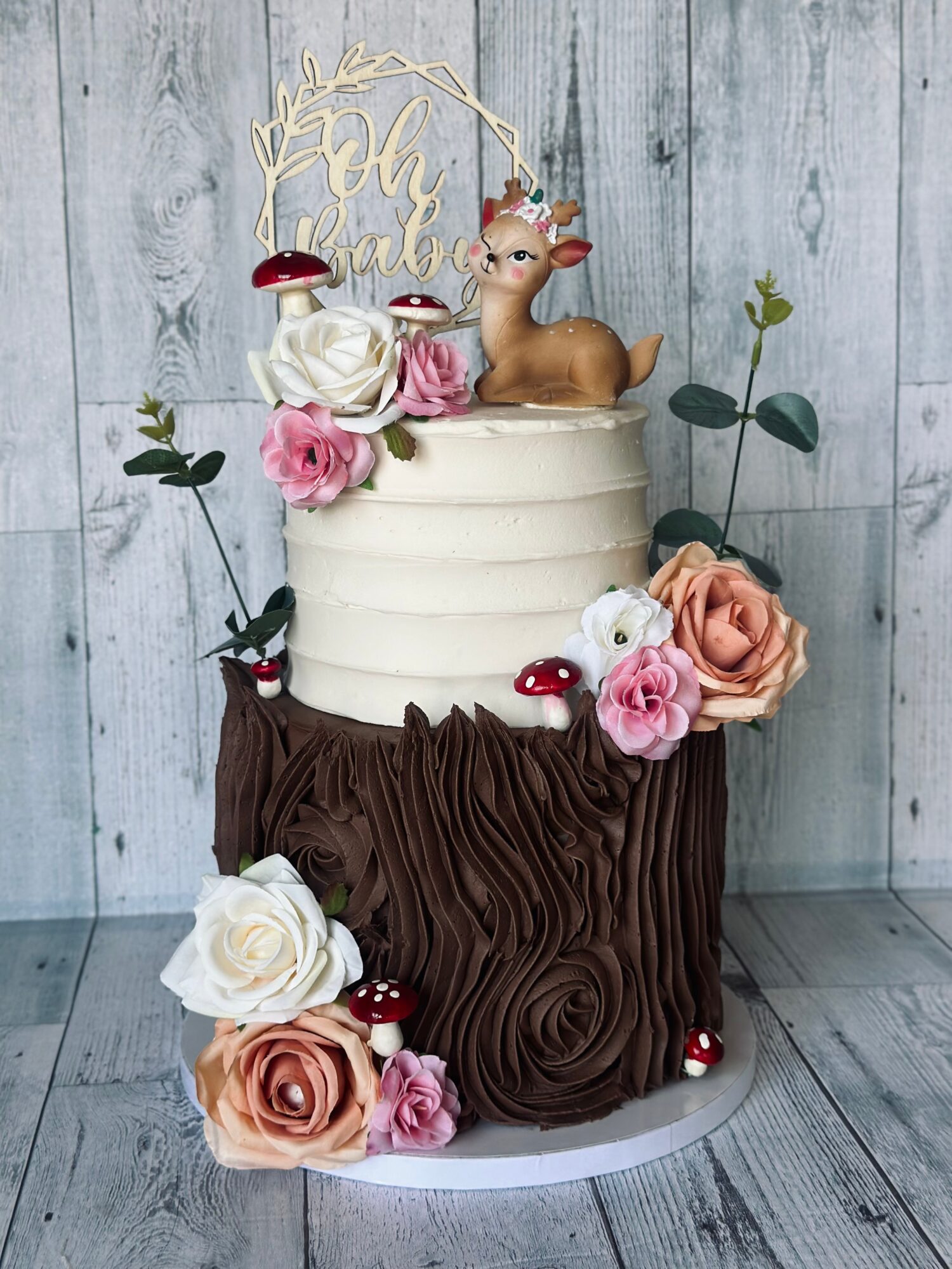 Two-tiered cake with a reindeer topper, decorated with pink and white roses, mushrooms, and greenery, against a wooden background.