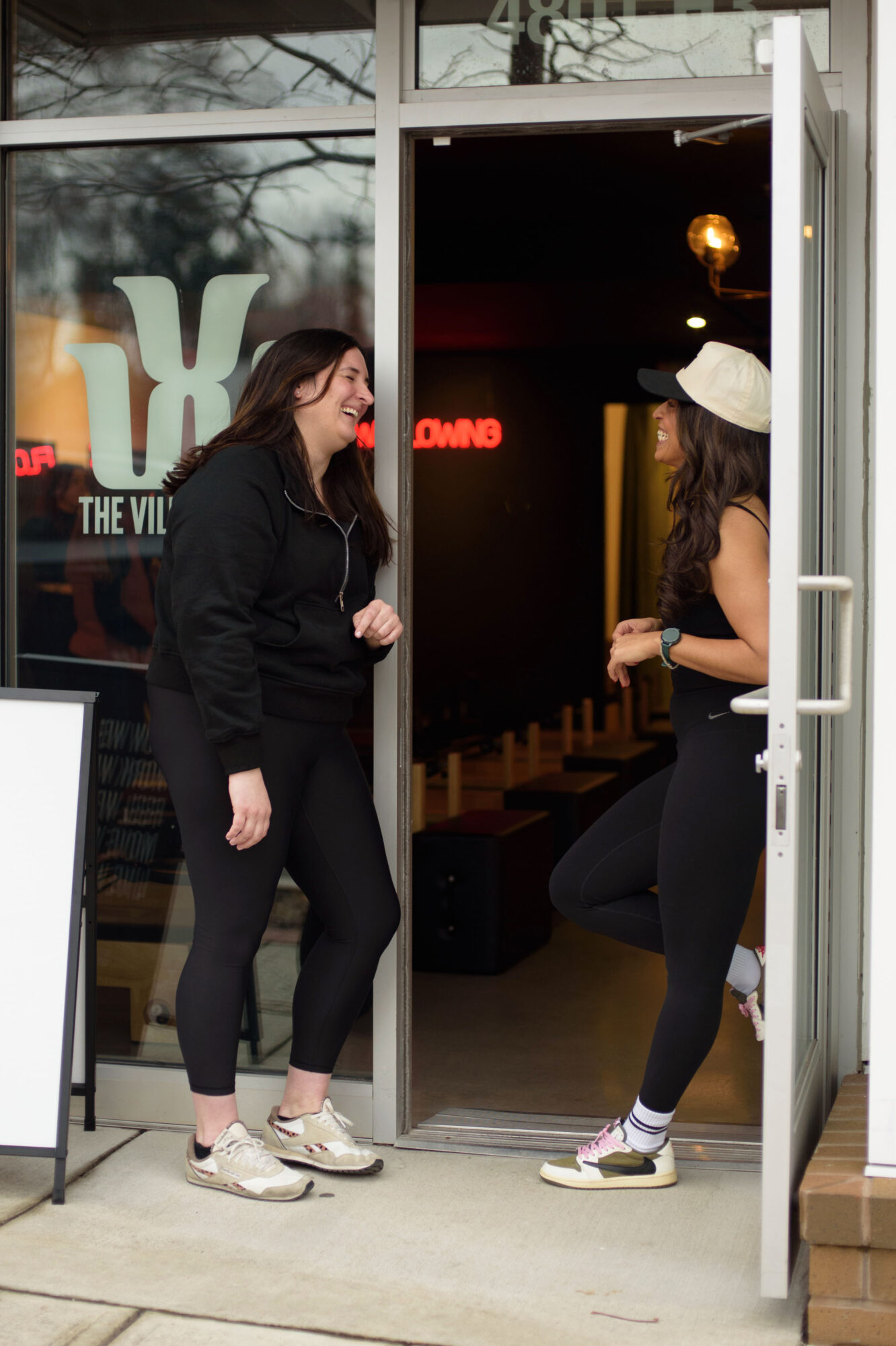 Two women stand and talk at the entrance of a building, one inside and one outside, with a glass door and window.