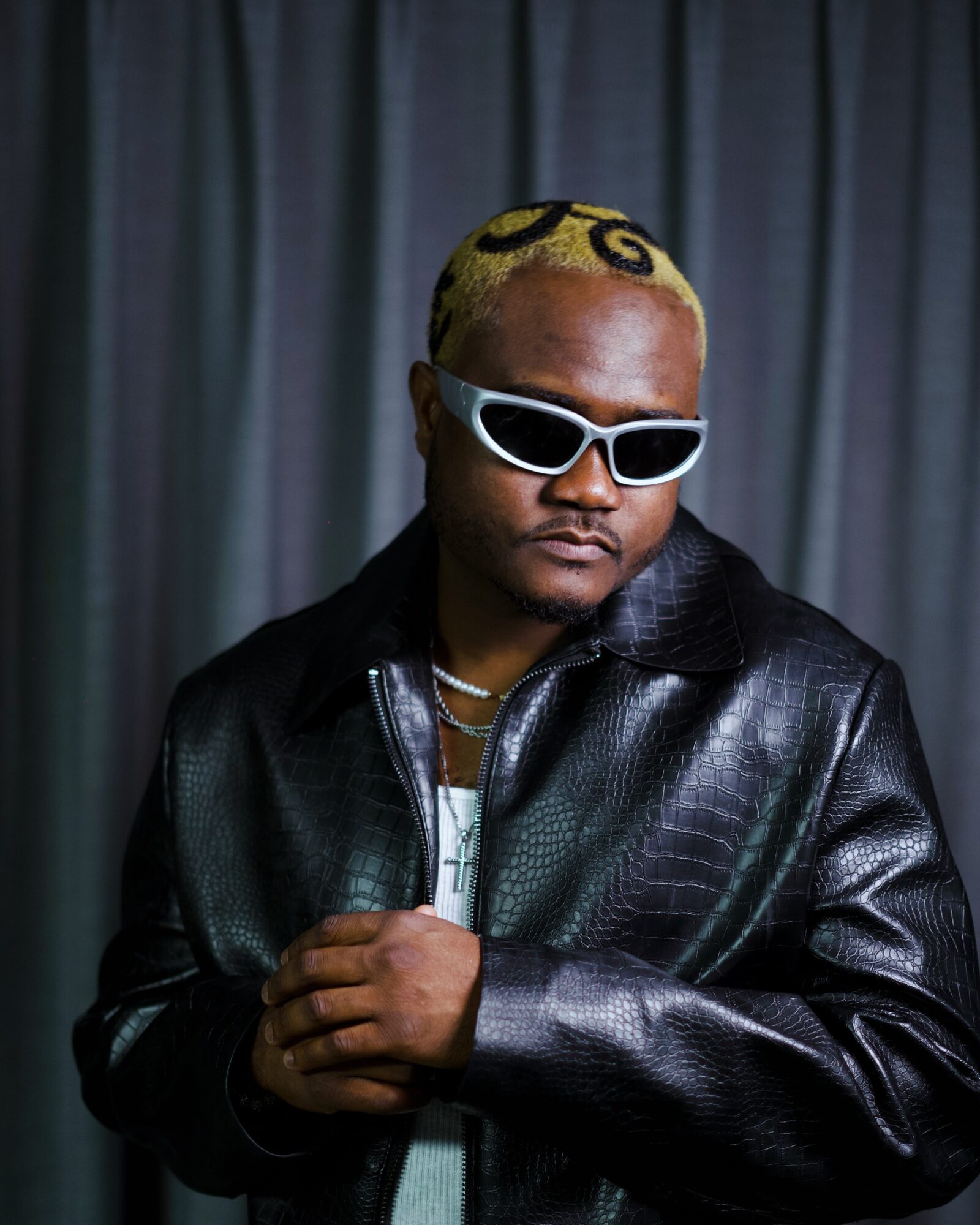 Person with yellow hair, wearing white sunglasses and a black leather jacket, standing in front of a dark curtain.
