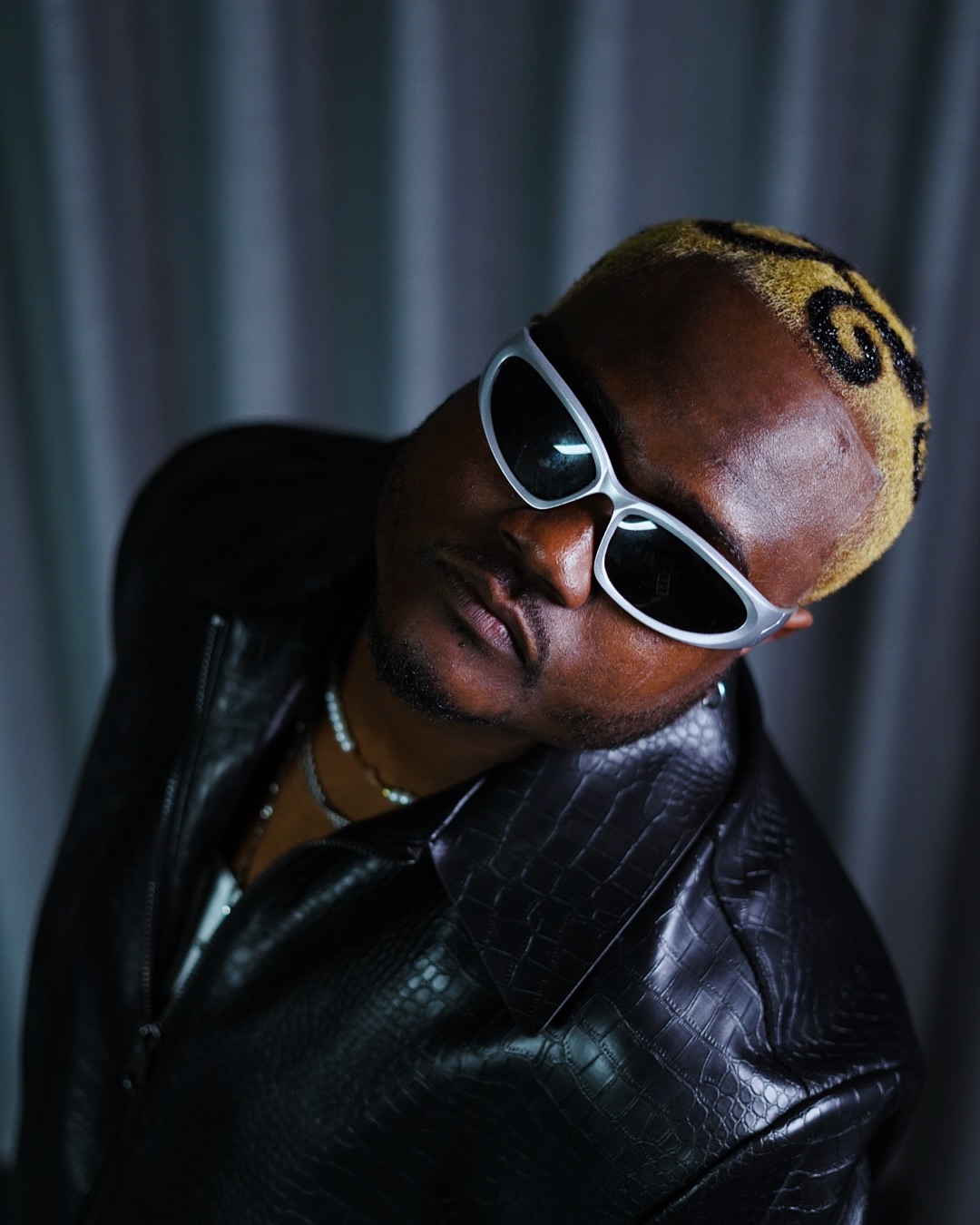 Person with blonde hair, wearing white sunglasses and a black leather jacket, tilting head to the side.