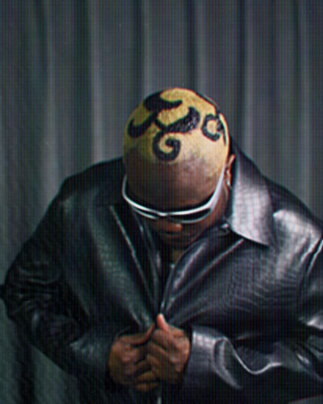 Person with a bald head and painted design, wearing sunglasses and a black leather jacket, looking downward.