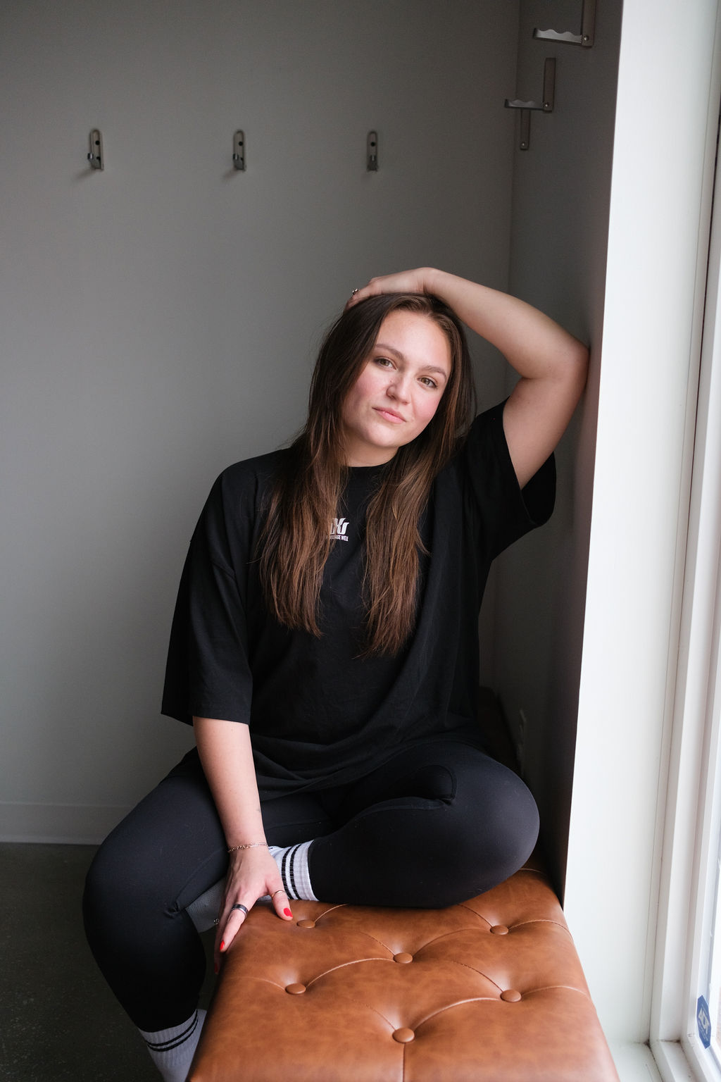 Young woman sitting on a brown cushioned stool near a window, with one hand on her head and a neutral expression.