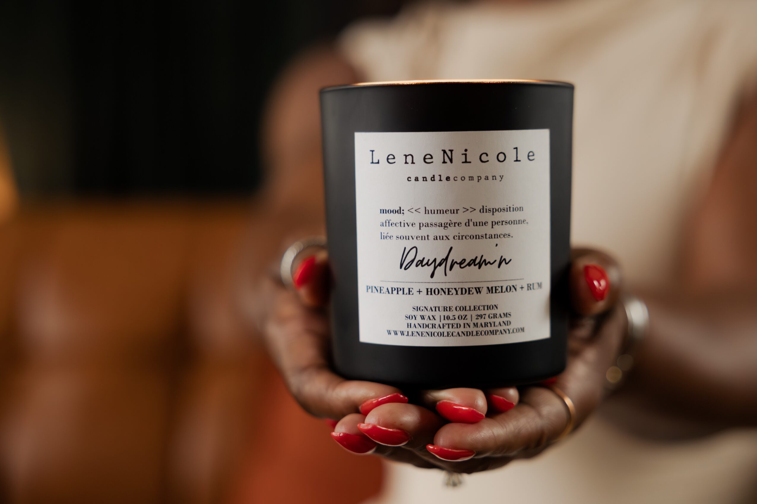 Person holding a black candle with a white label, hands with red nail polish, blurred background.