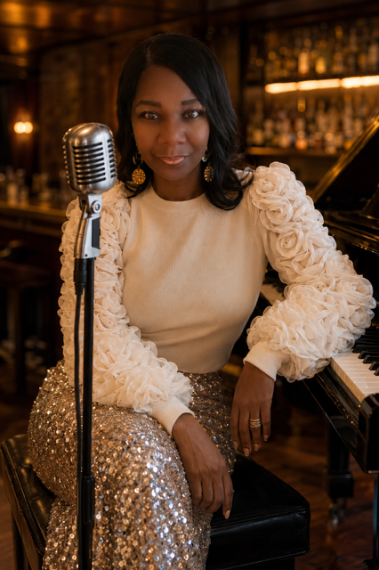 Woman sitting at a piano with a vintage microphone, wearing a cream top with ruffled sleeves and a sequined skirt, in a dimly lit setting.