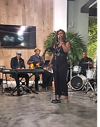 A woman singing into a microphone with a band playing instruments behind her indoors, with a large plant and a TV screen nearby.