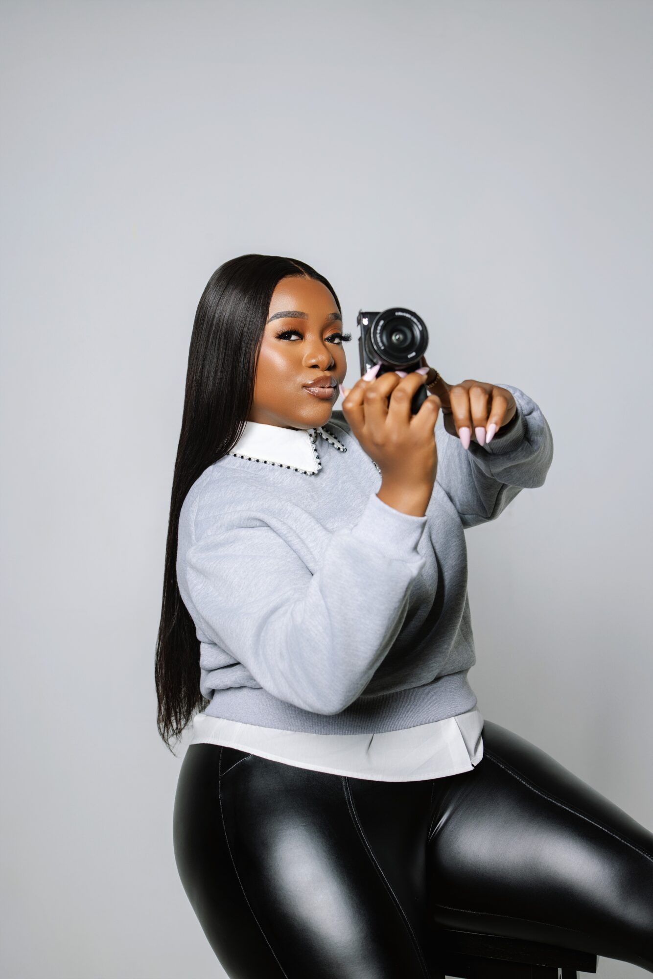 Woman with long black hair holding a camera, wearing a gray sweatshirt and black shiny pants, sitting against a plain background.