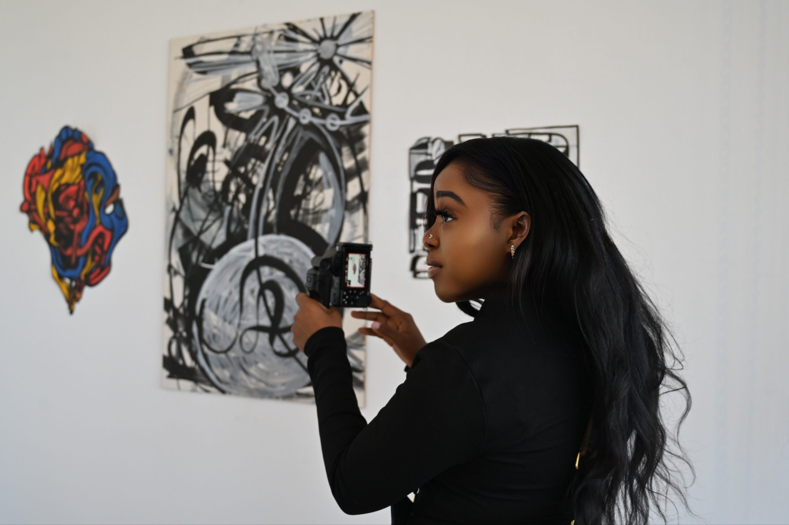 Woman with long black hair taking a photo of artwork on gallery wall, black shirt, side profile, white background