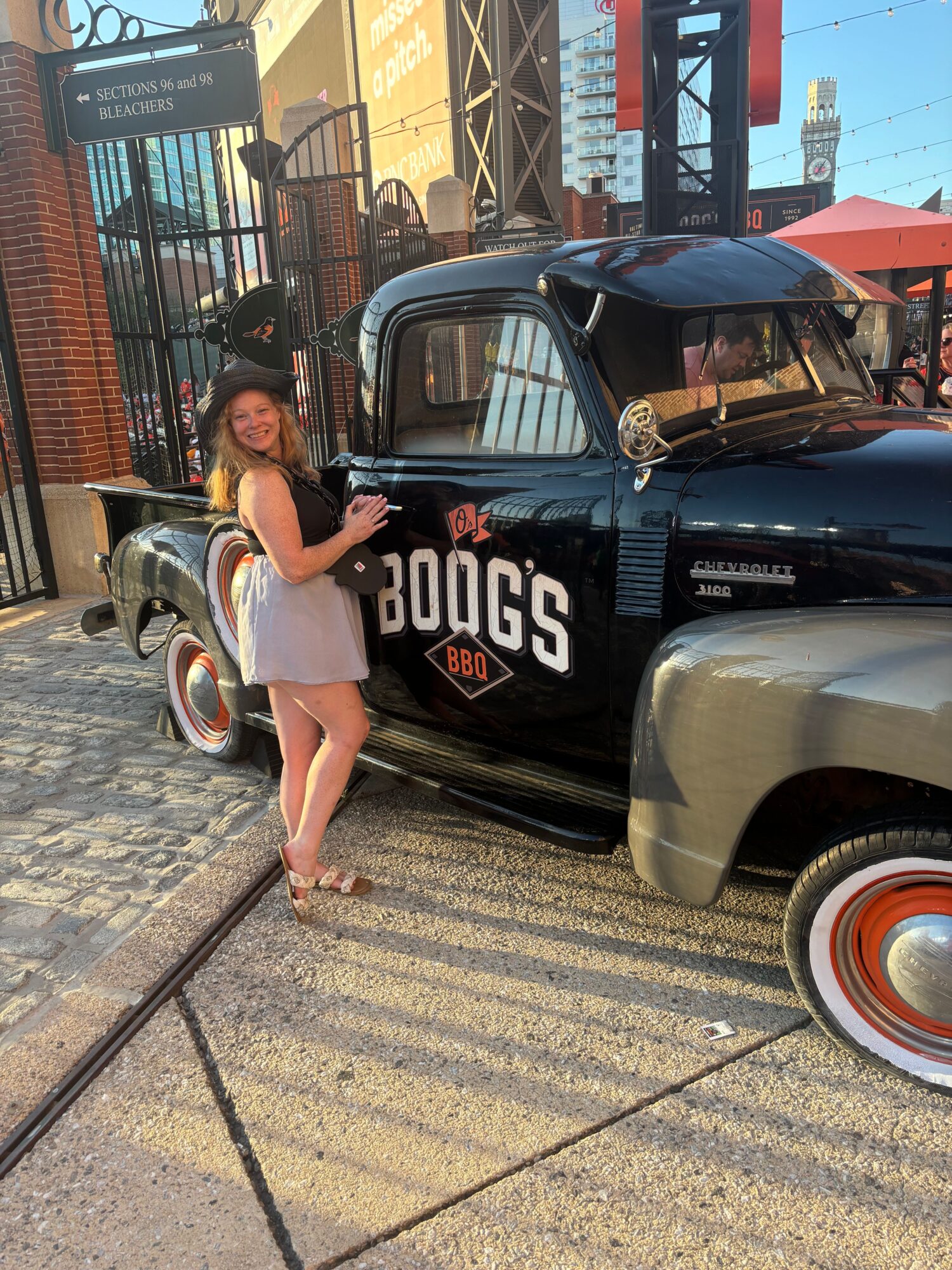 Woman standing next to a vintage car on a city street, holding a bag, with buildings and a fence in the background.