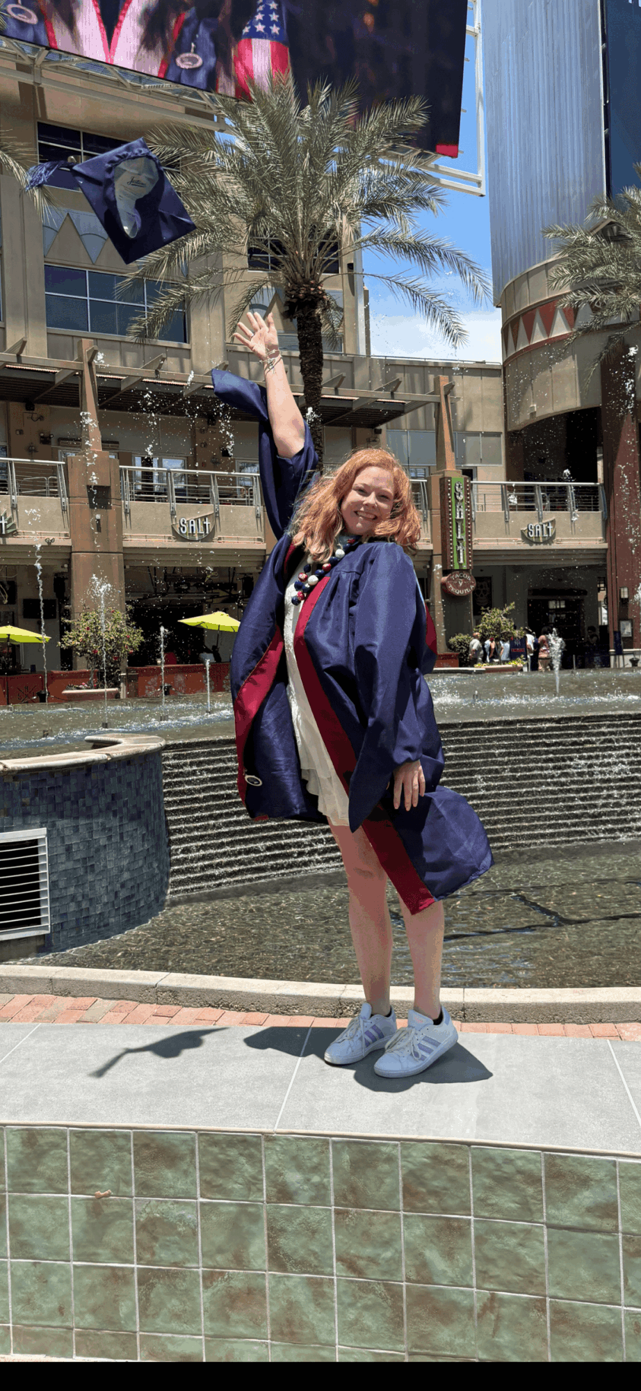 Person with long hair in a graduation gown standing outdoors near a fountain, holding a cap in the air.
