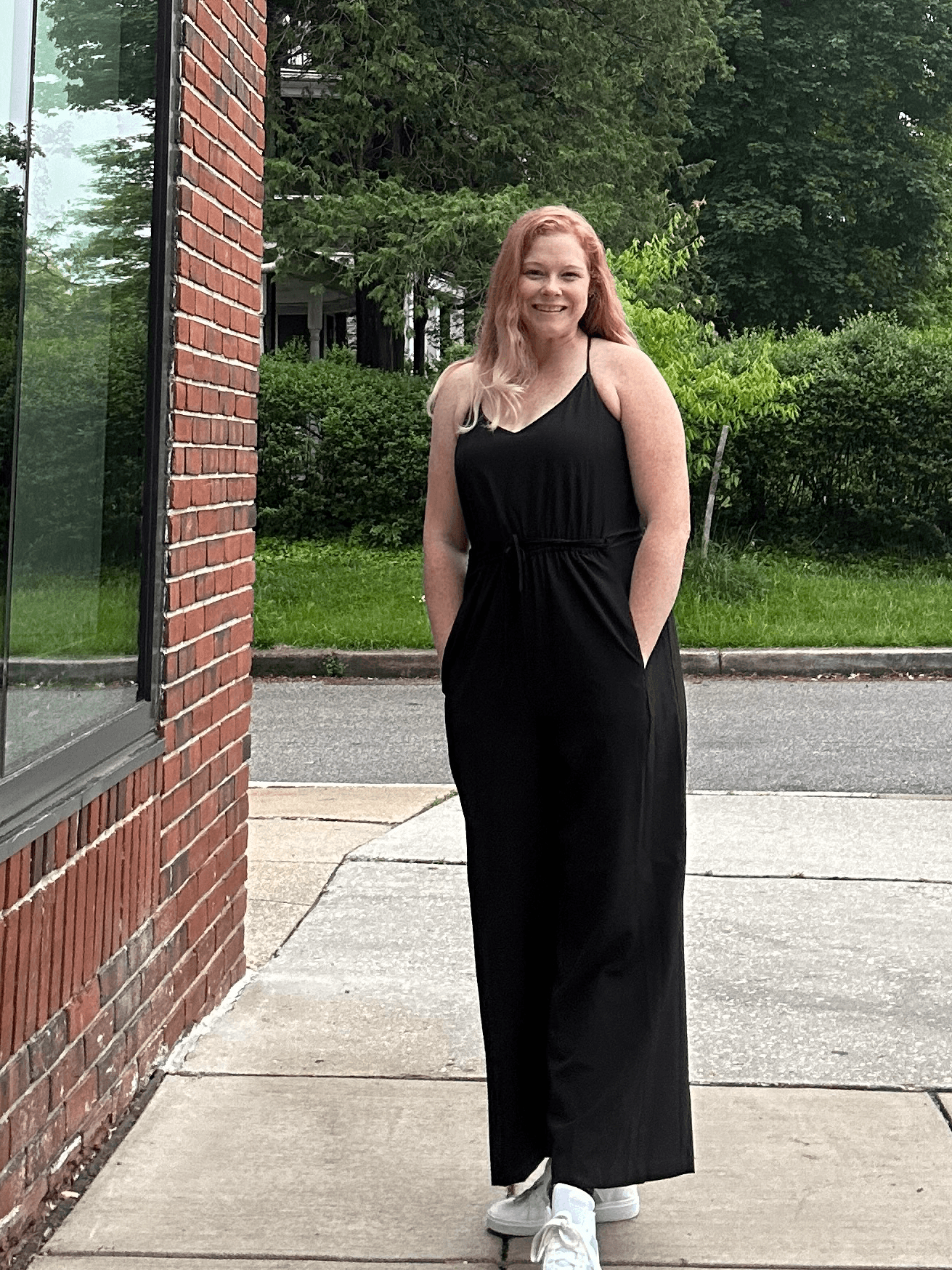 Woman in black jumpsuit standing on sidewalk near brick building, smiling, with trees and grass in background.