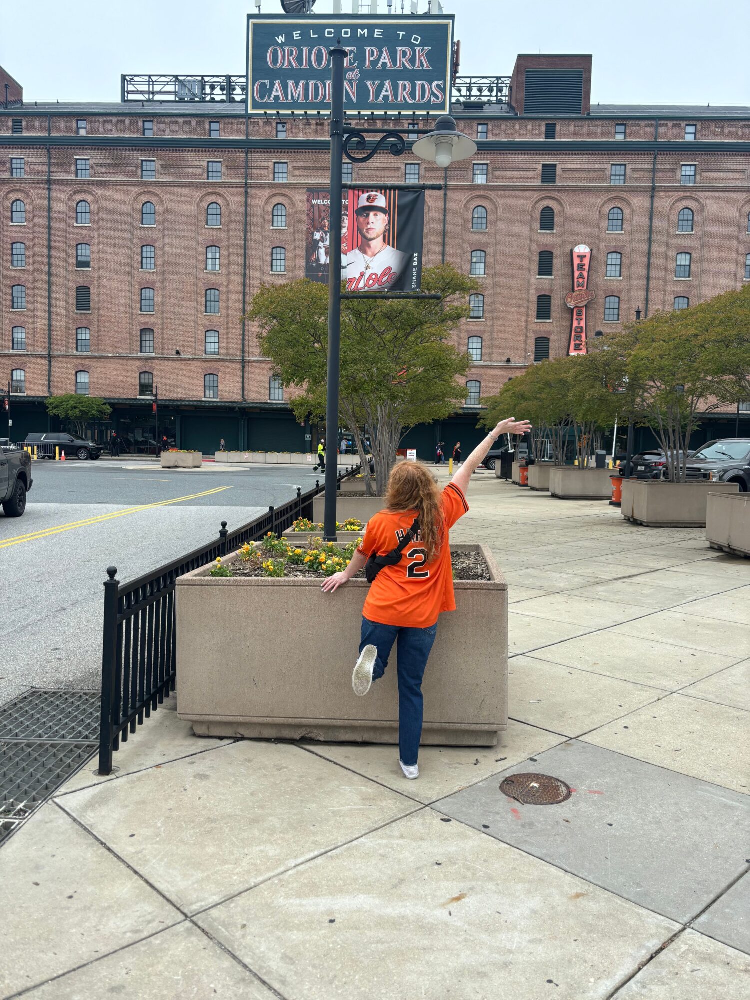 Person in orange jacket and dark pants pointing at a sign on a city sidewalk, with buildings in the background.