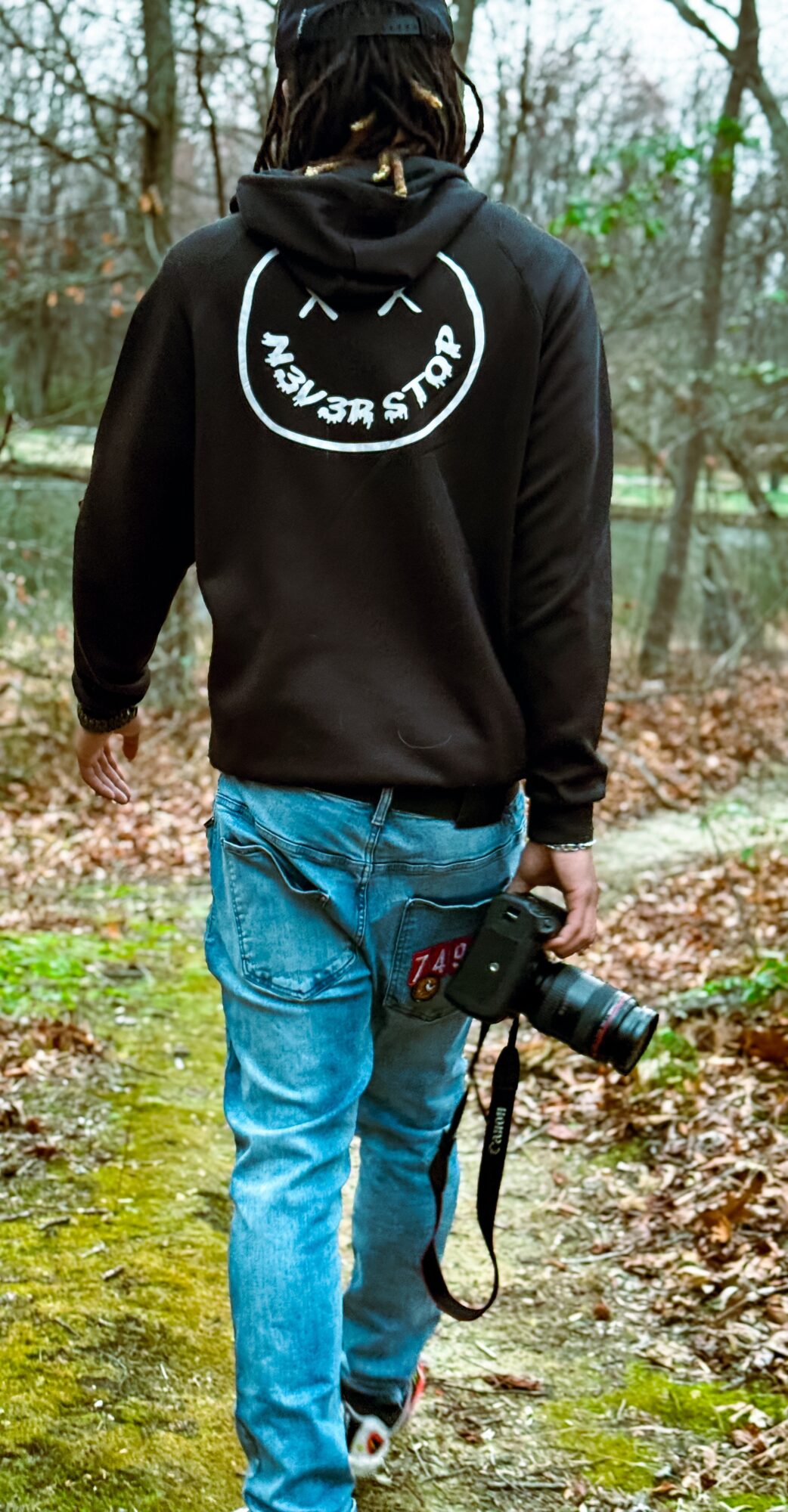 Person walking in a wooded area holding a camera, wearing a black hoodie and blue jeans.