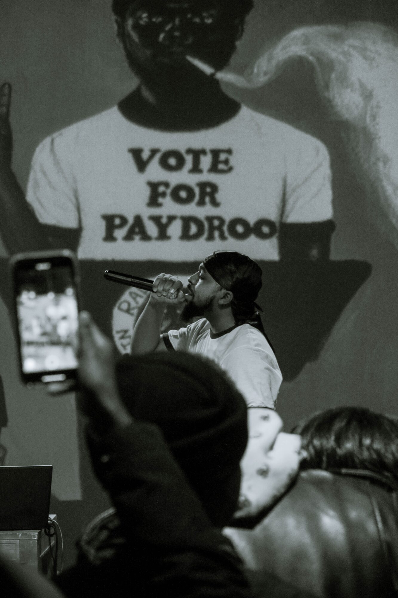 Person speaking into a microphone at a rally, with a large screen behind showing a person wearing a 'VOTE FOR PAYDROO' T-shirt, audience members in foreground.
