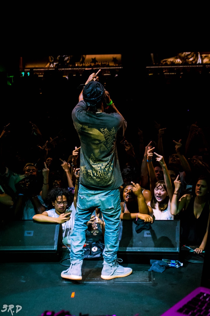 Performer on stage facing audience, with crowd reaching out, in a dark setting with stage lighting, people taking photos.