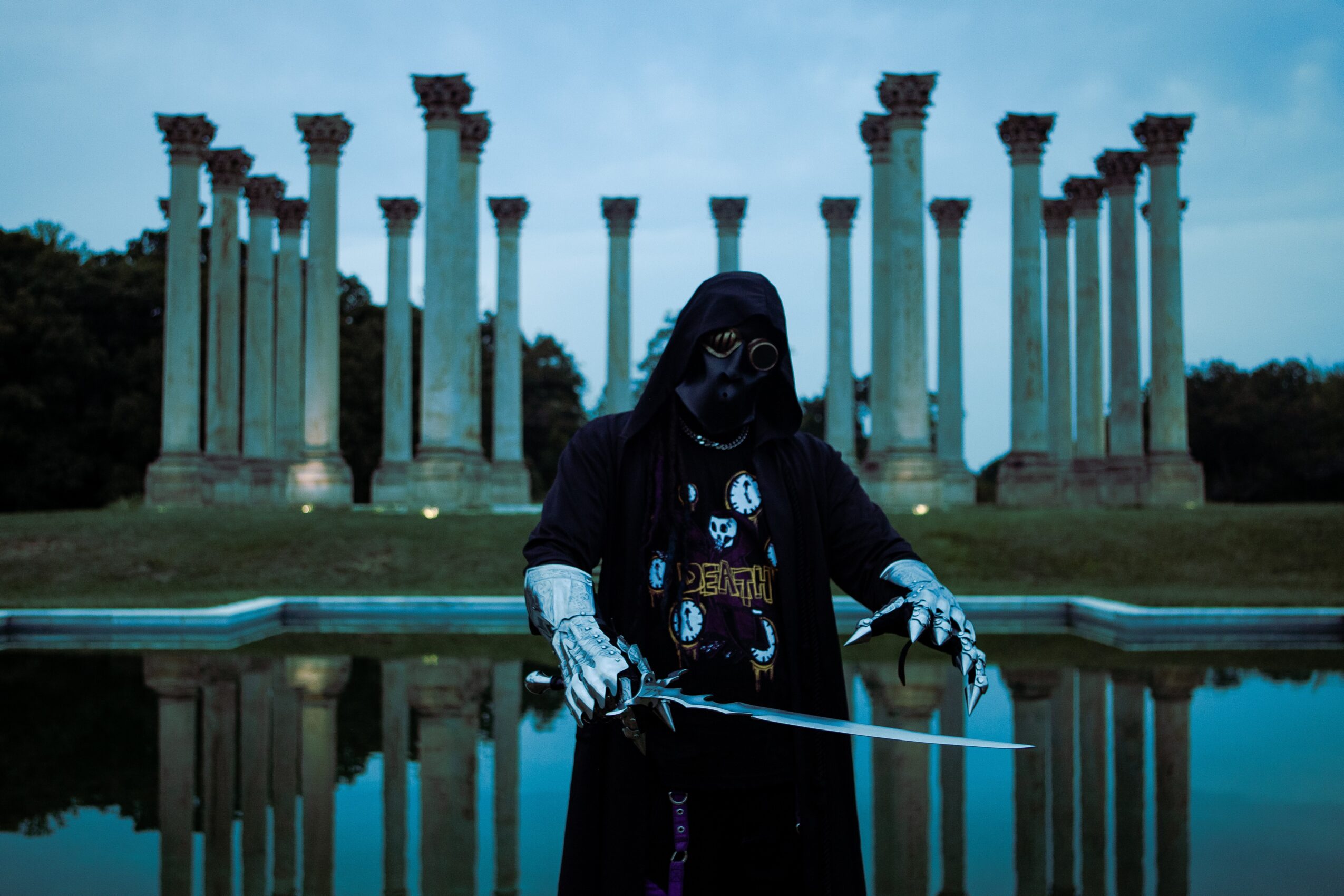 Person dressed in dark clothing and mask, holding a sword, standing in front of columns and water reflection.