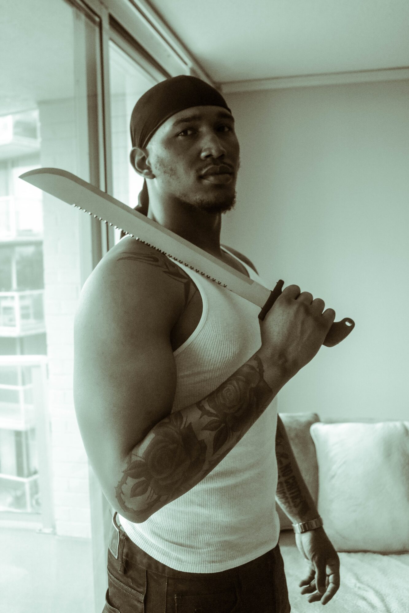 Young man with a knife on his shoulder, wearing a white tank top and a black headband, standing indoors near a window.
