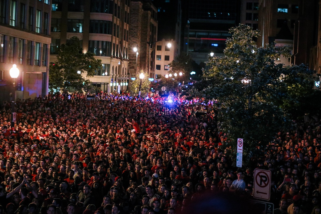 Large crowd gathered outdoors at night in an urban area with tall buildings and trees, illuminated by streetlights and city lights.