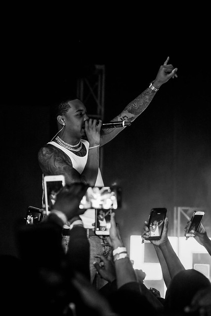 Performer with tattoos and jewelry sings into microphone, raising one hand, audience capturing with phones, dark background.