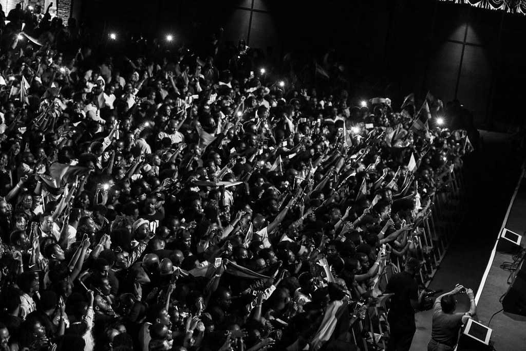 Crowd of people at a concert or event, many raising hands, in black and white.
