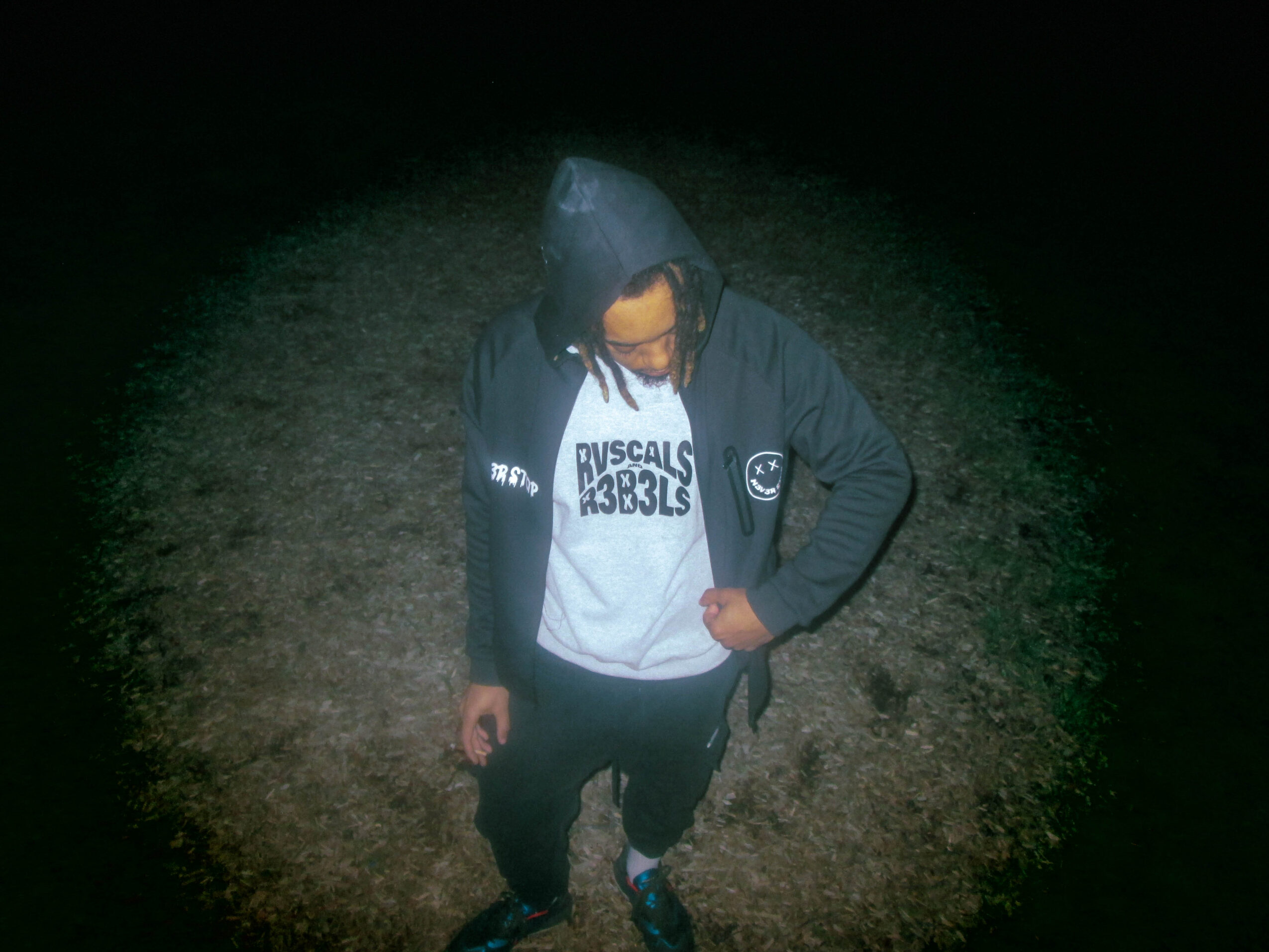Person wearing a hoodie and jacket, standing outdoors at night, looking down, with dark background.