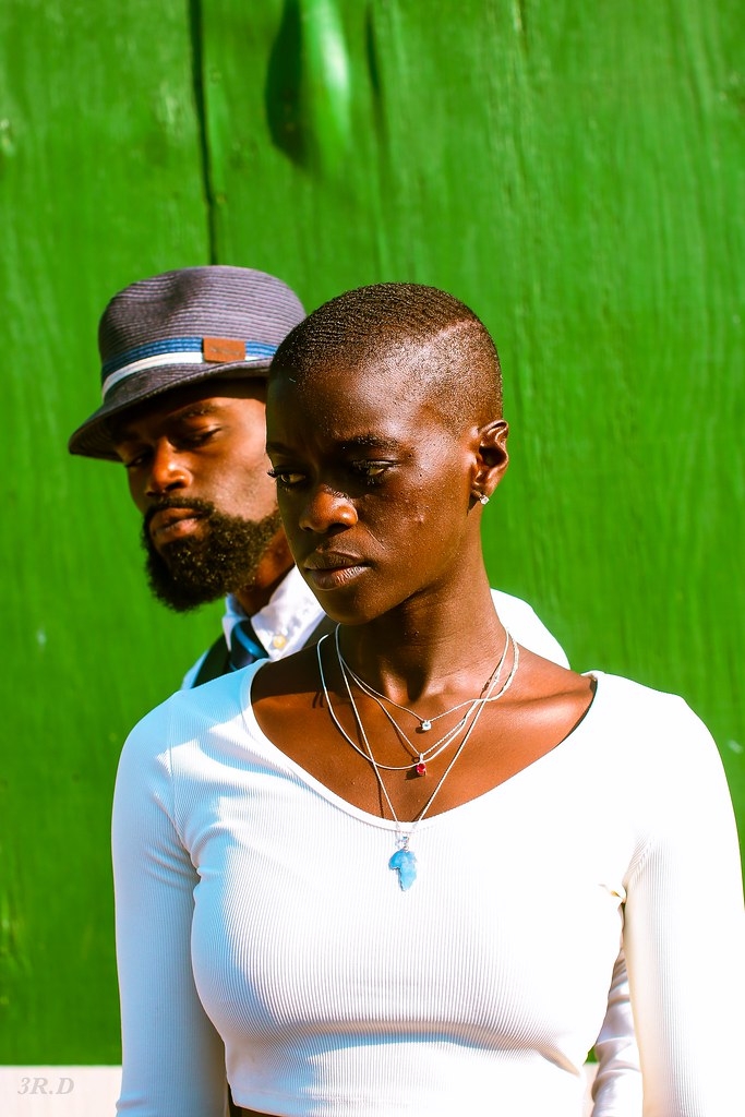 Two people, a woman in the foreground and a man in the background, stand against a green background. The woman has short hair and wears a white top with layered necklaces. The man wears a hat and has a beard.