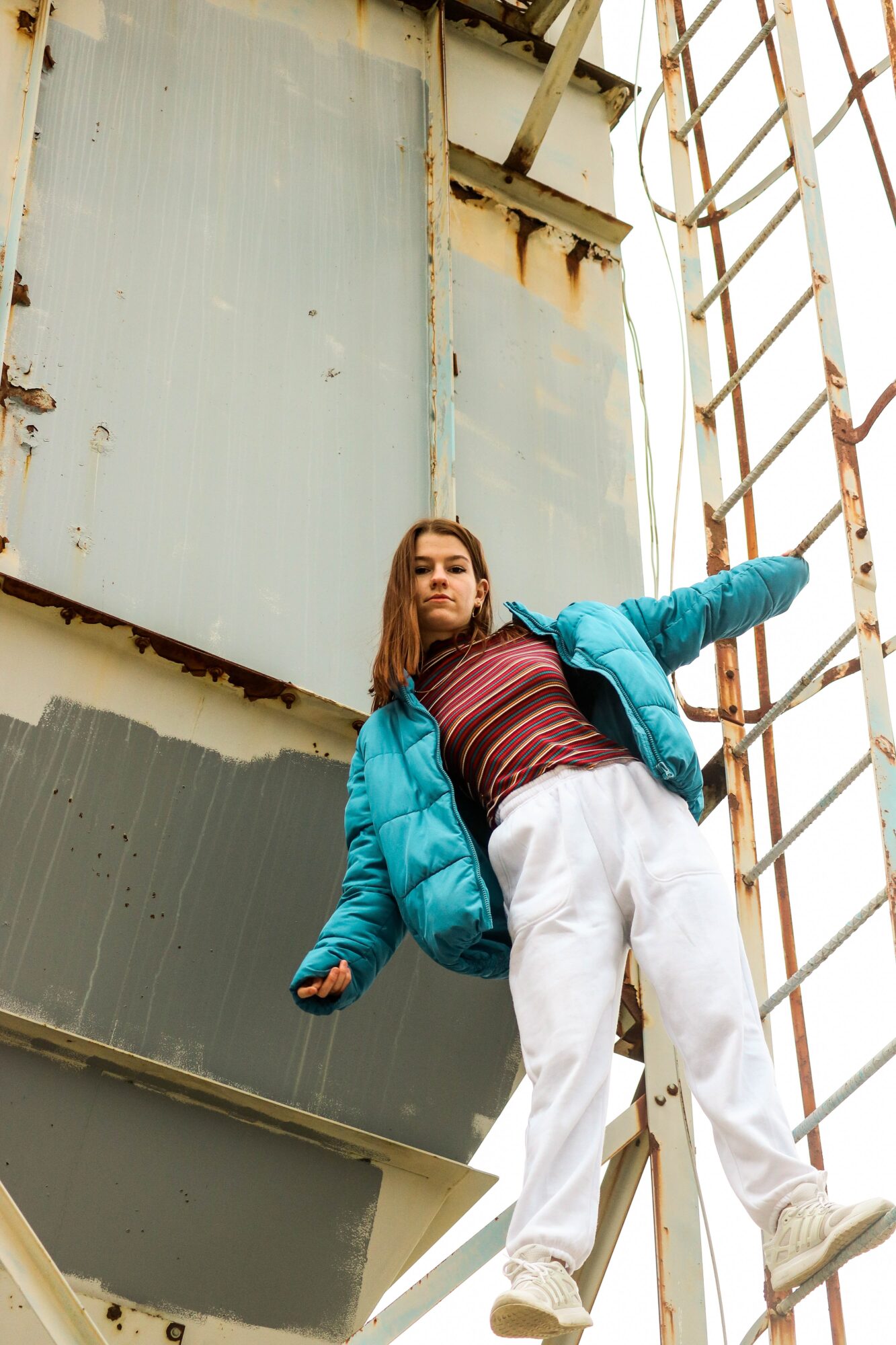 Young woman in a blue jacket and white pants leaning against a rusted industrial structure with metal stairs.