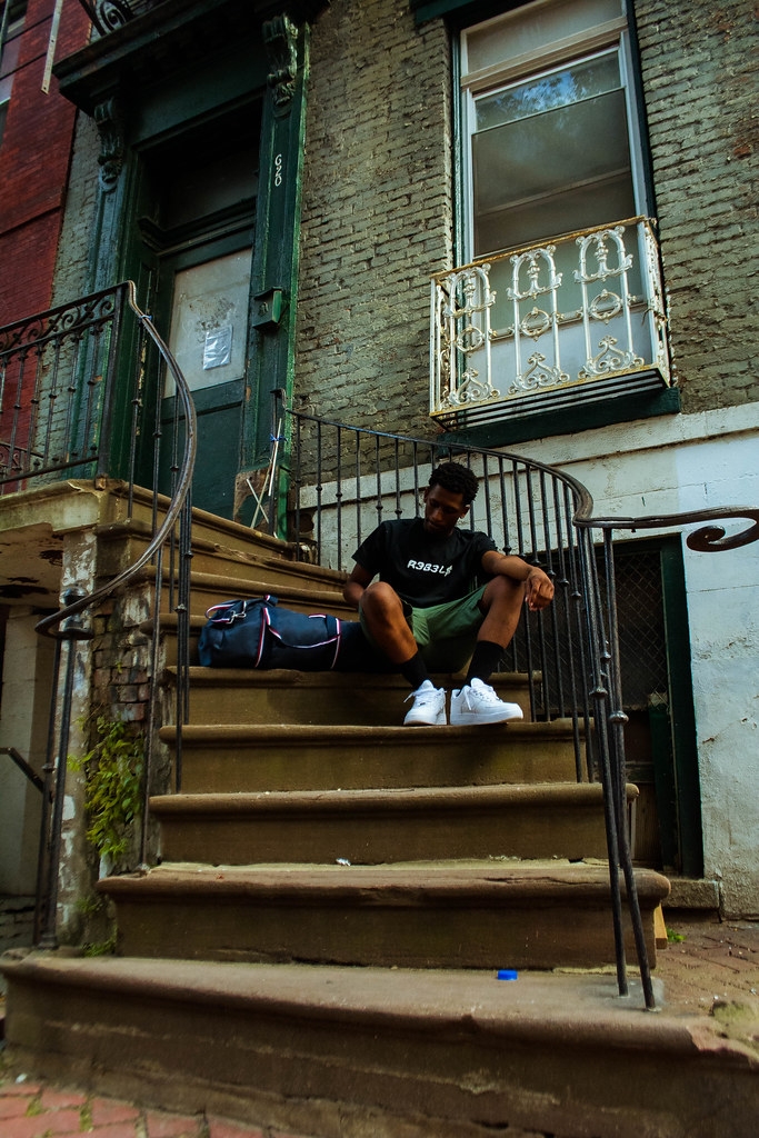 Person sitting on front steps of a brick building, with a backpack beside them, wearing a black shirt and white shoes.
