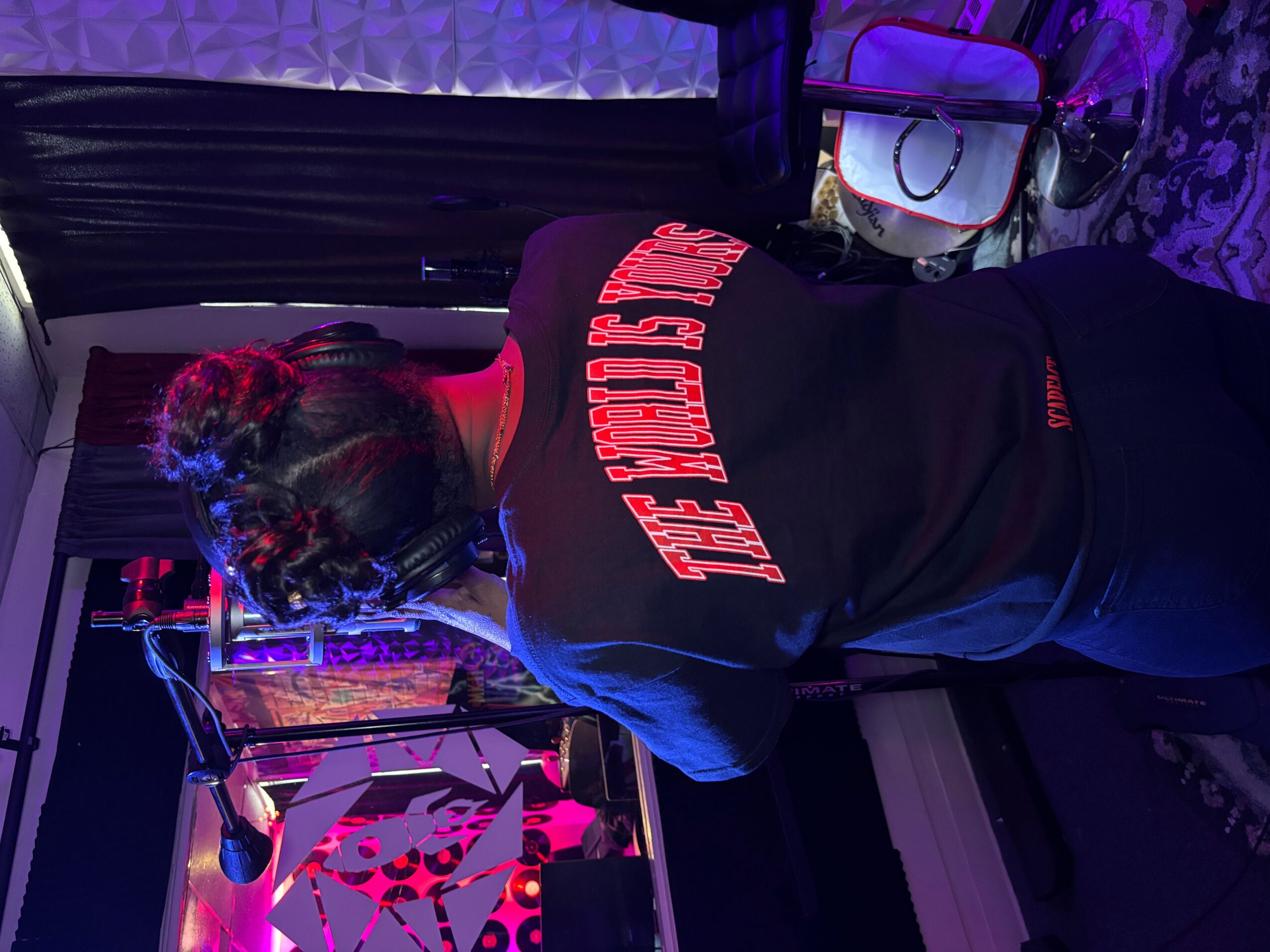 Person with braided hair wearing a black shirt with red text, standing in a room with purple lighting and electronic equipment.
