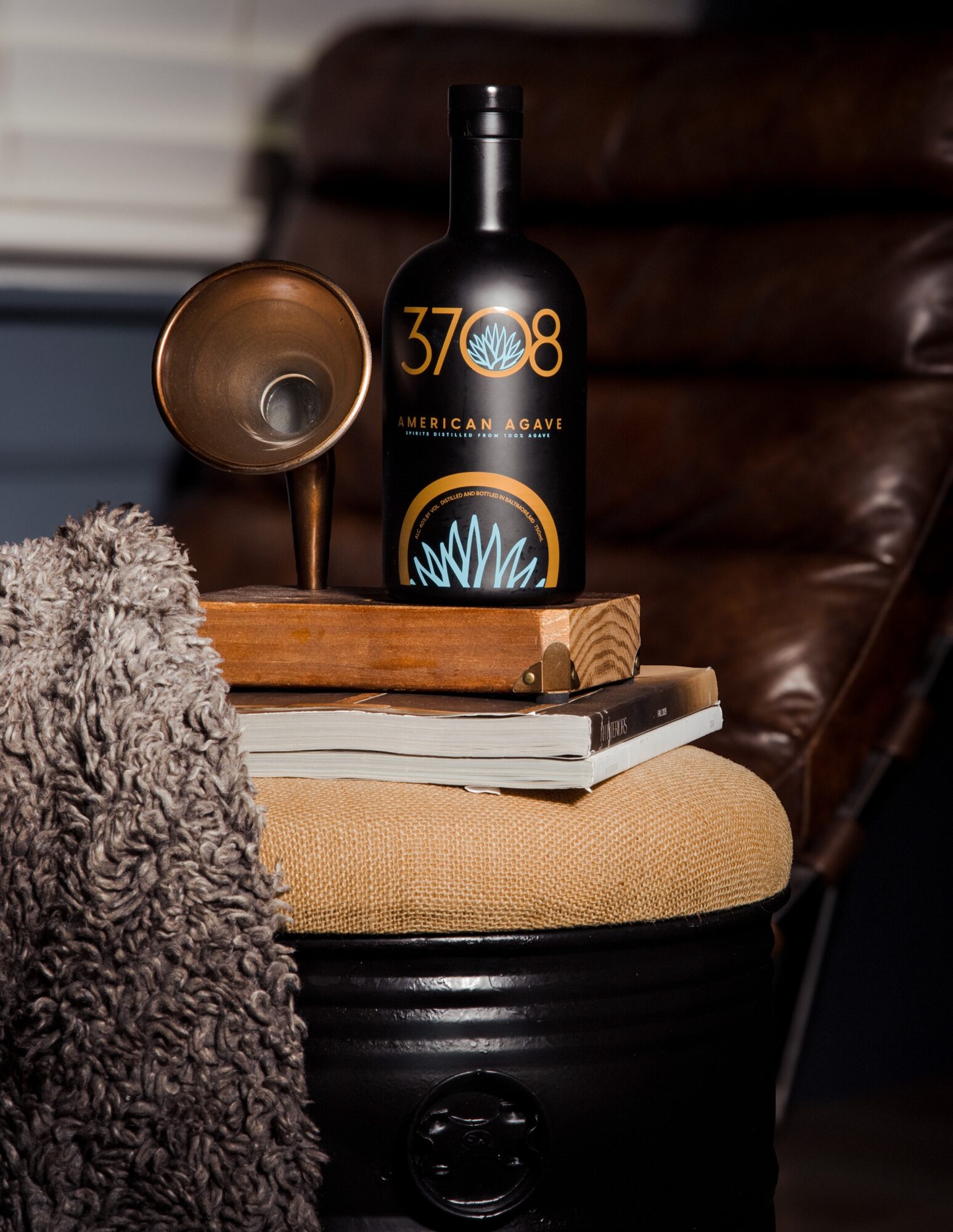 Bottle of alcohol, vintage speaker, and books on a wooden tray on a cushioned stool, with a leather chair in background.