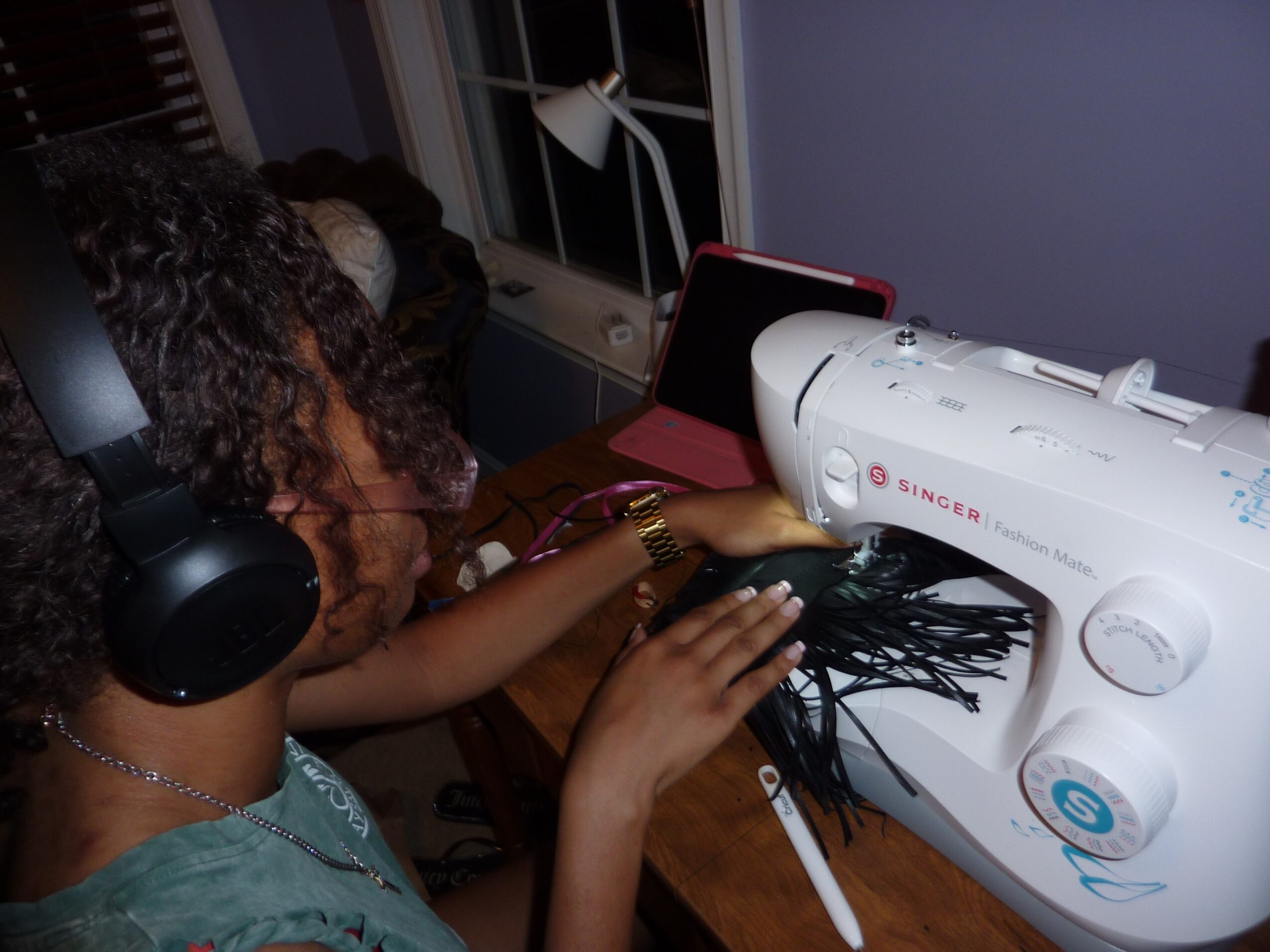 Person sewing black fabric with a sewing machine, wearing headphones, in a room with a window and a pink chair.
