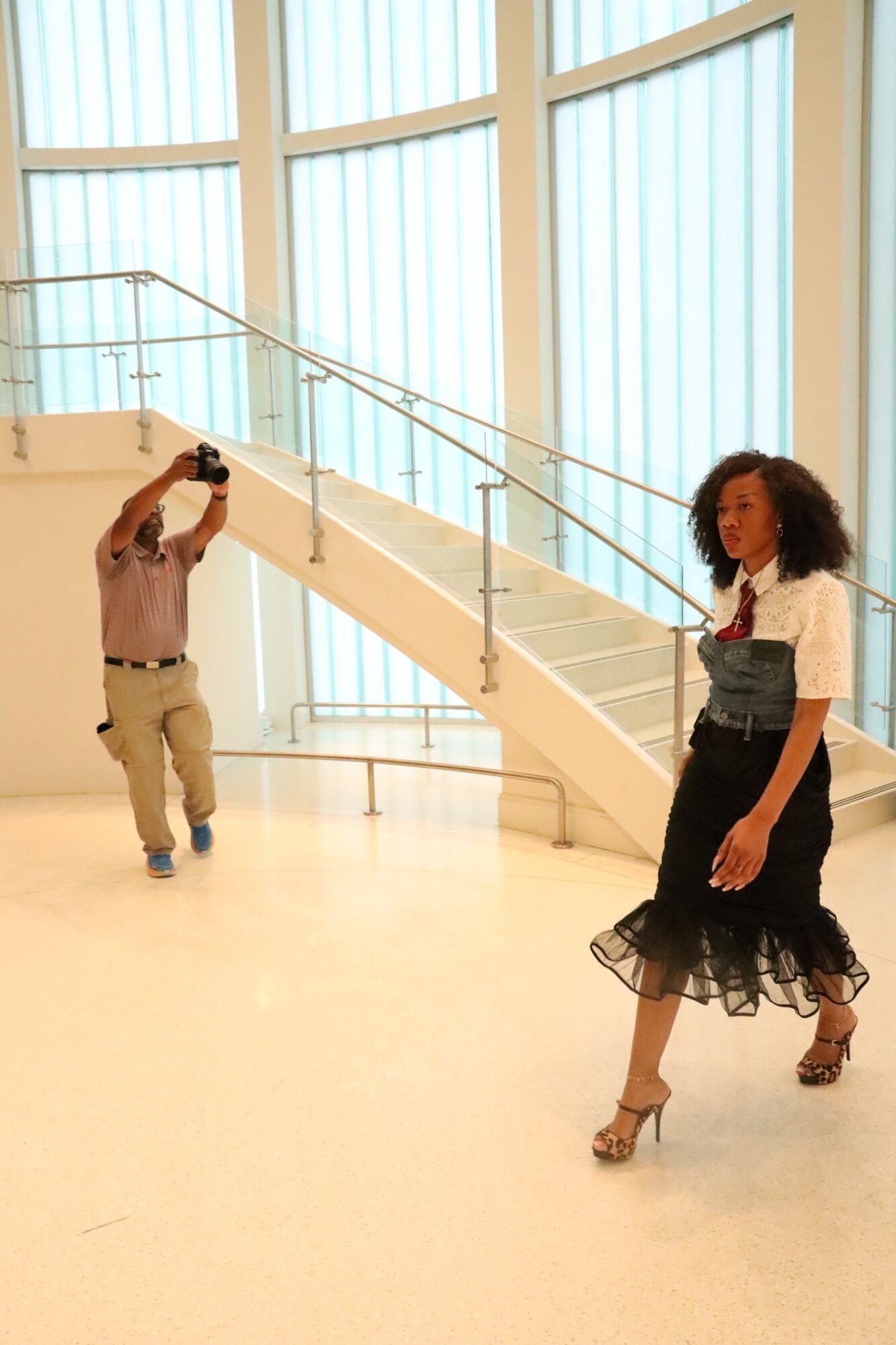 Two women in a gallery with large windows, one taking a photo, the other standing near a sculpture, both dressed stylishly.
