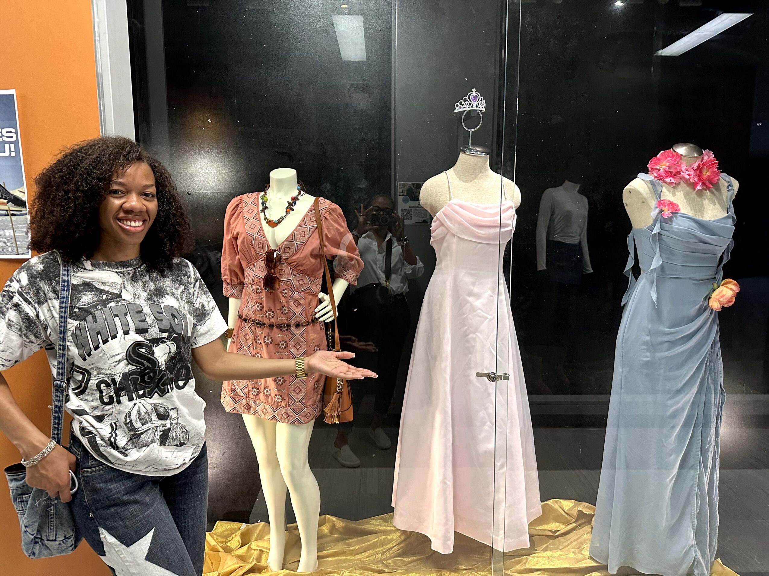 Woman smiling and pointing at mannequins dressed in elegant gowns and accessories, display case behind them, gold-colored floor, indoor setting.