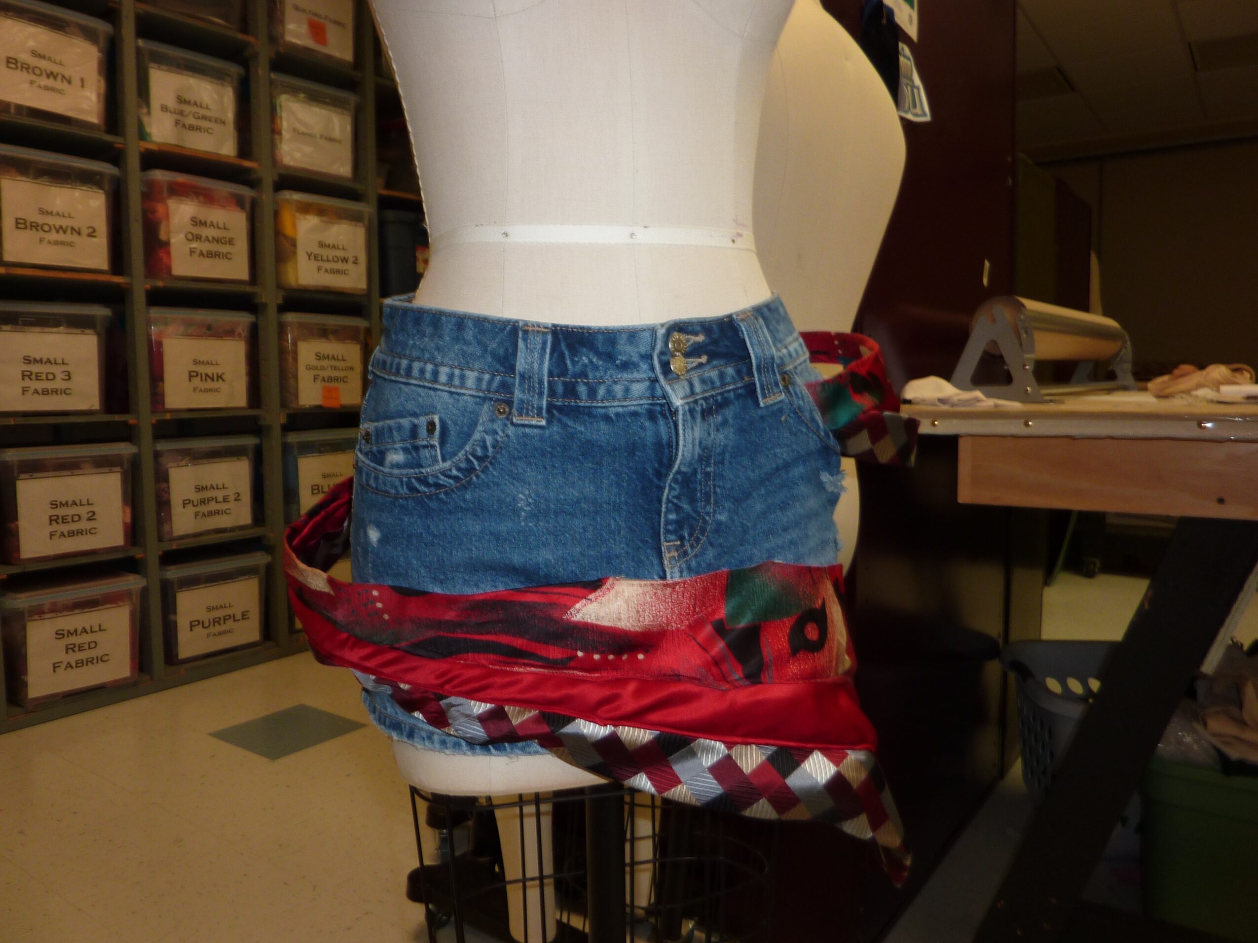 Mannequin torso wearing jeans and a white top, with a red and black patterned cloth draped around the waist, in a room with storage drawers and a table.