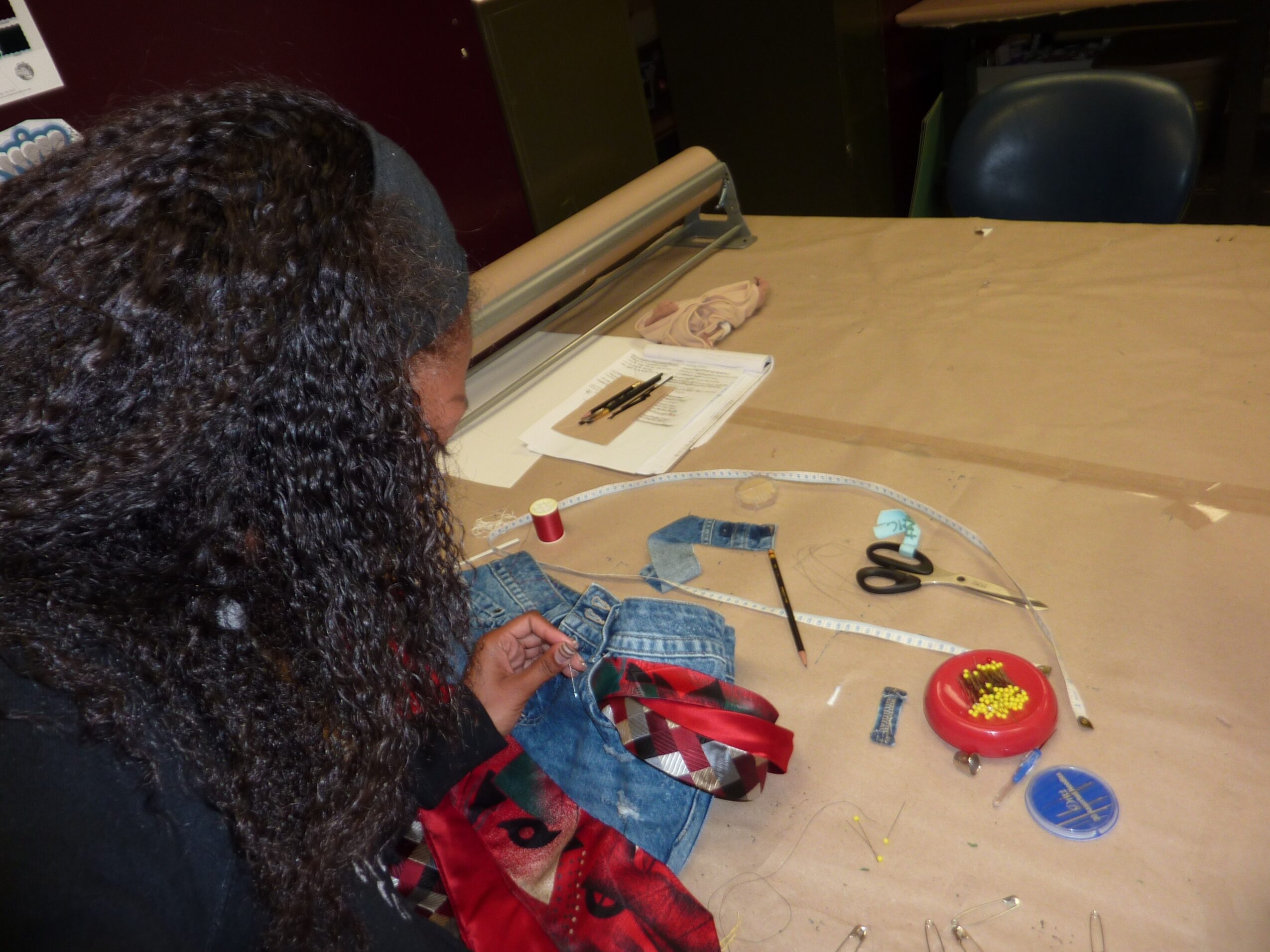 Person with curly hair working on a sewing project at a table with sewing tools and fabric, viewed from the side.
