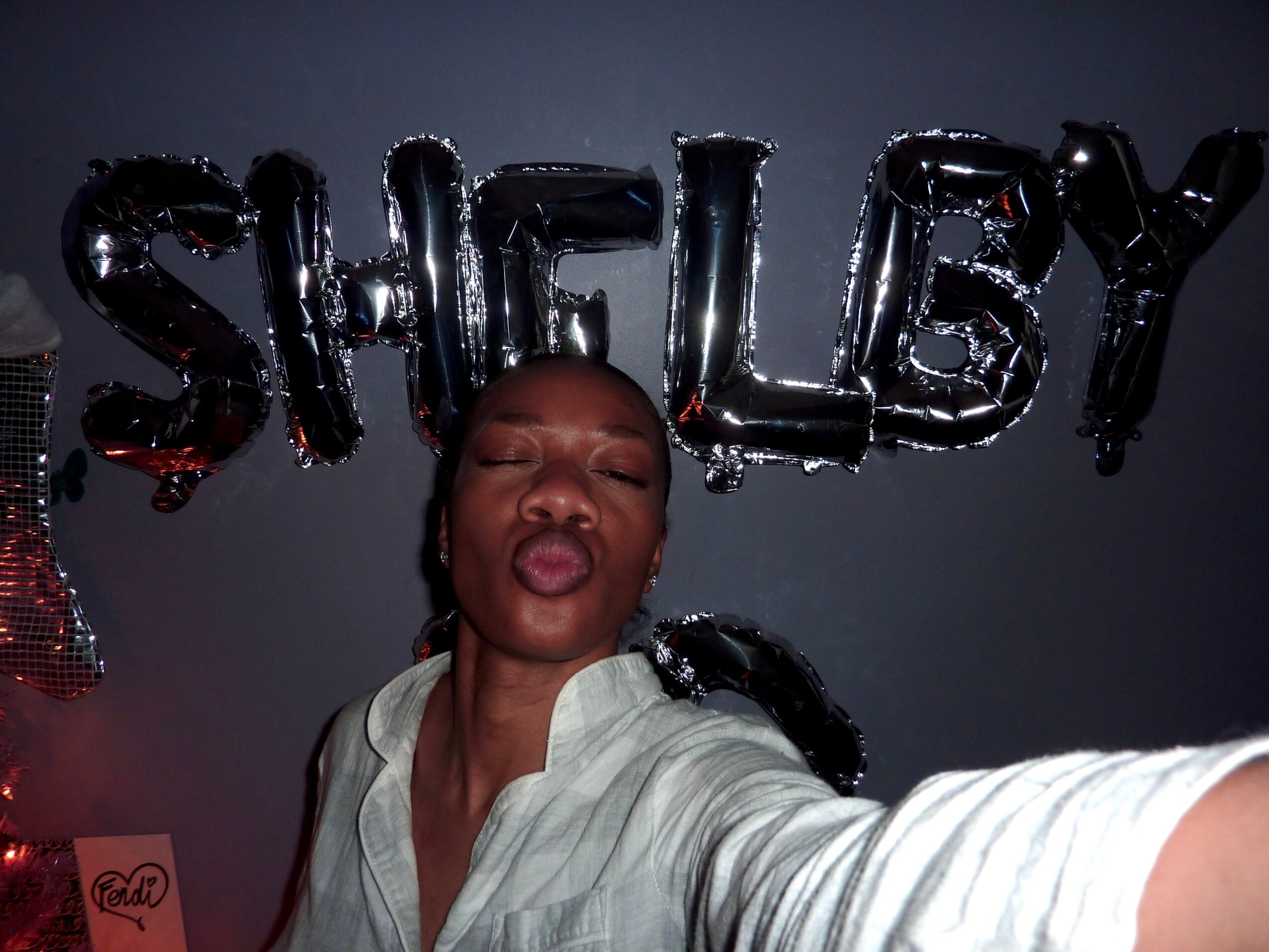 Person making a kiss face in front of shiny balloon letters spelling 'SHELBY'.