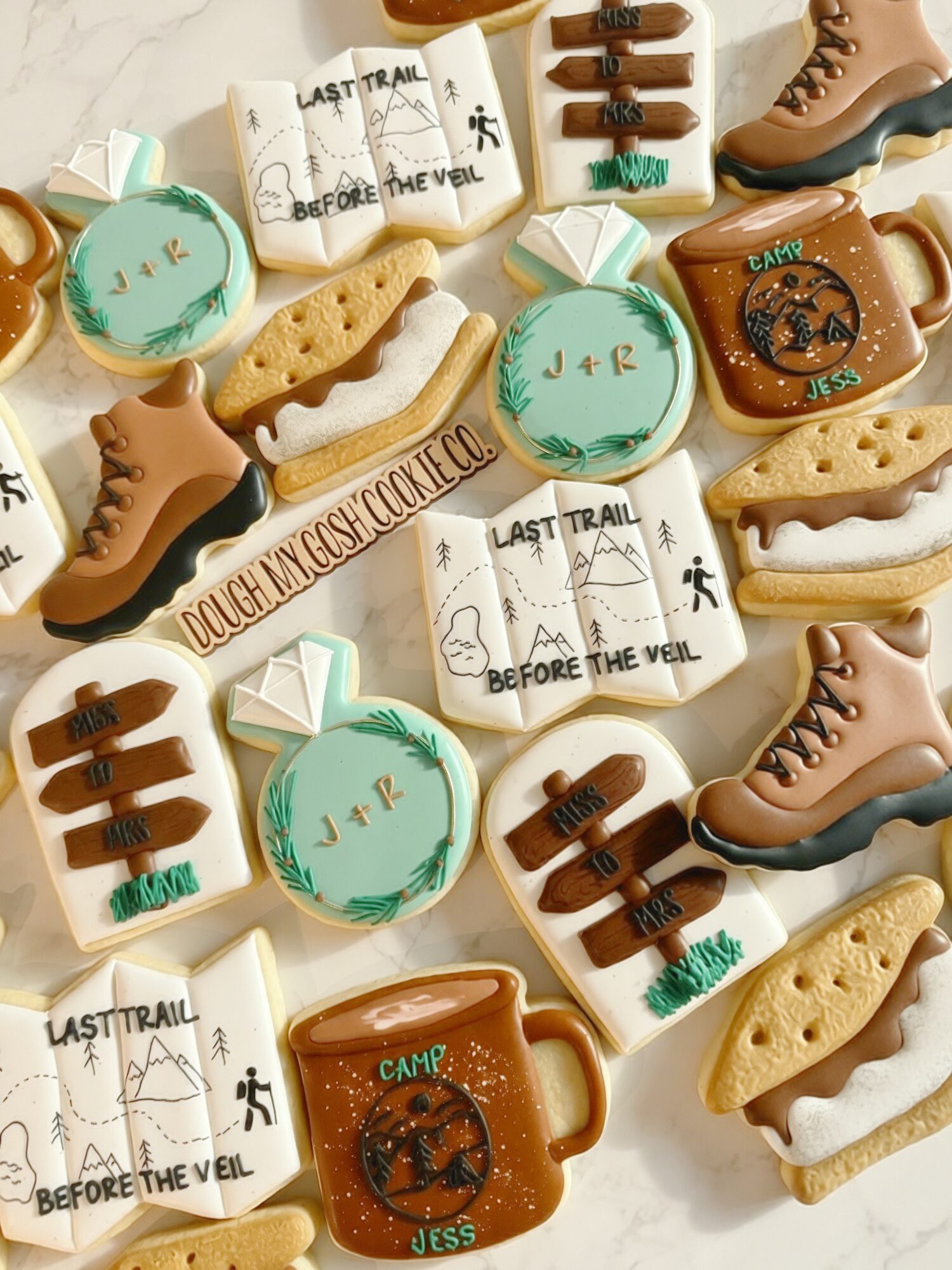 Assorted cookies shaped like boots, hats, and signs with outdoor and trail themes, decorated with icing and edible images.