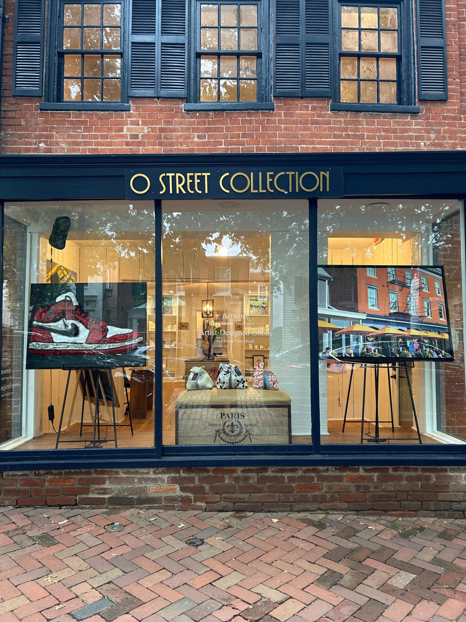 Storefront with large glass window displaying clothing and a red and white vehicle inside, brick sidewalk outside.