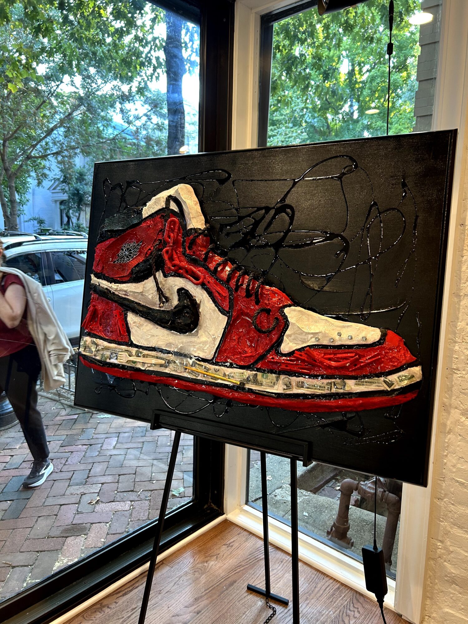 Painting of a red, white, and black sneaker with a Nike logo on a black background, displayed indoors near a window.