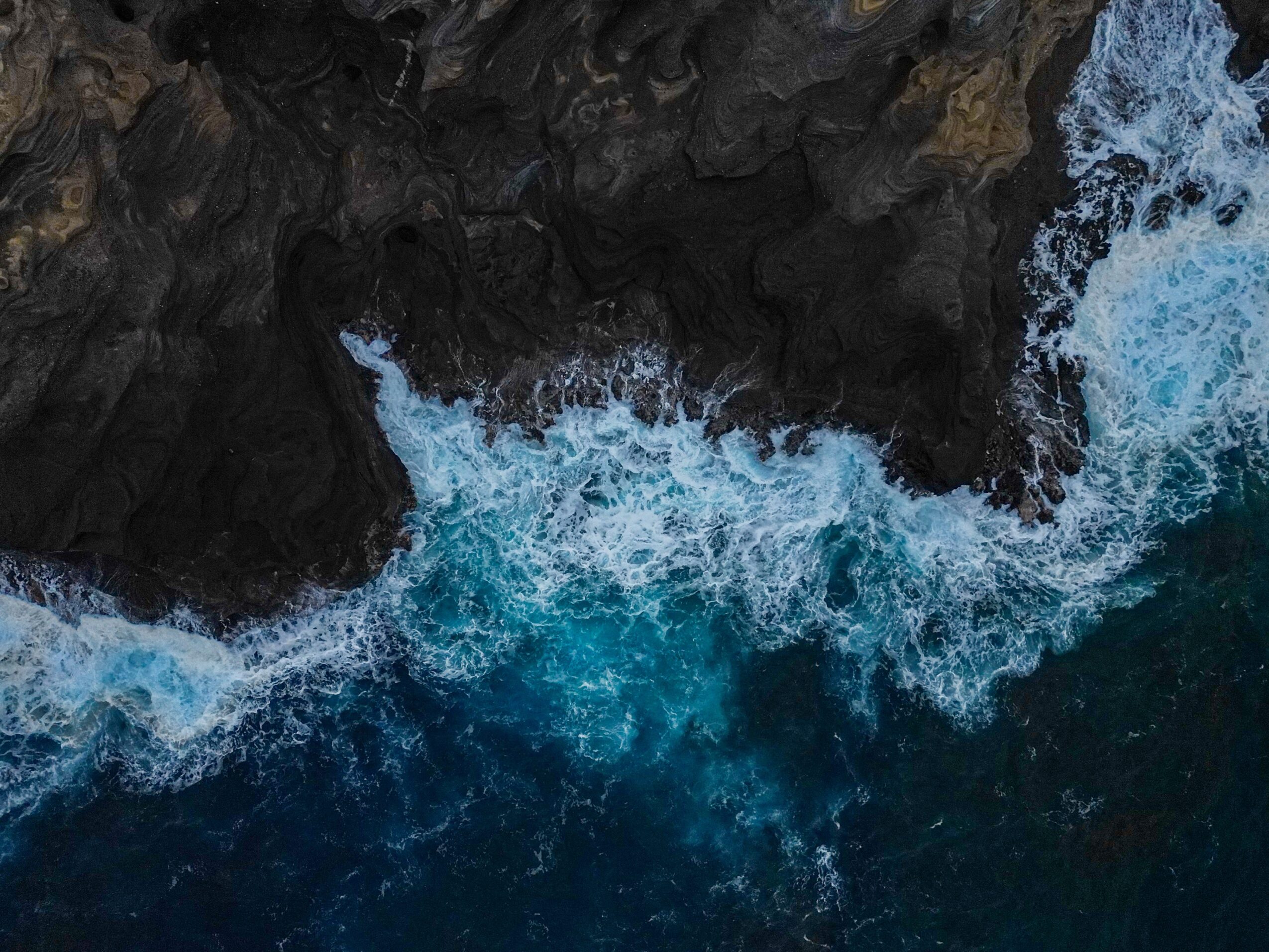 Aerial view of dark rocky coastline with blue ocean waves crashing against it.